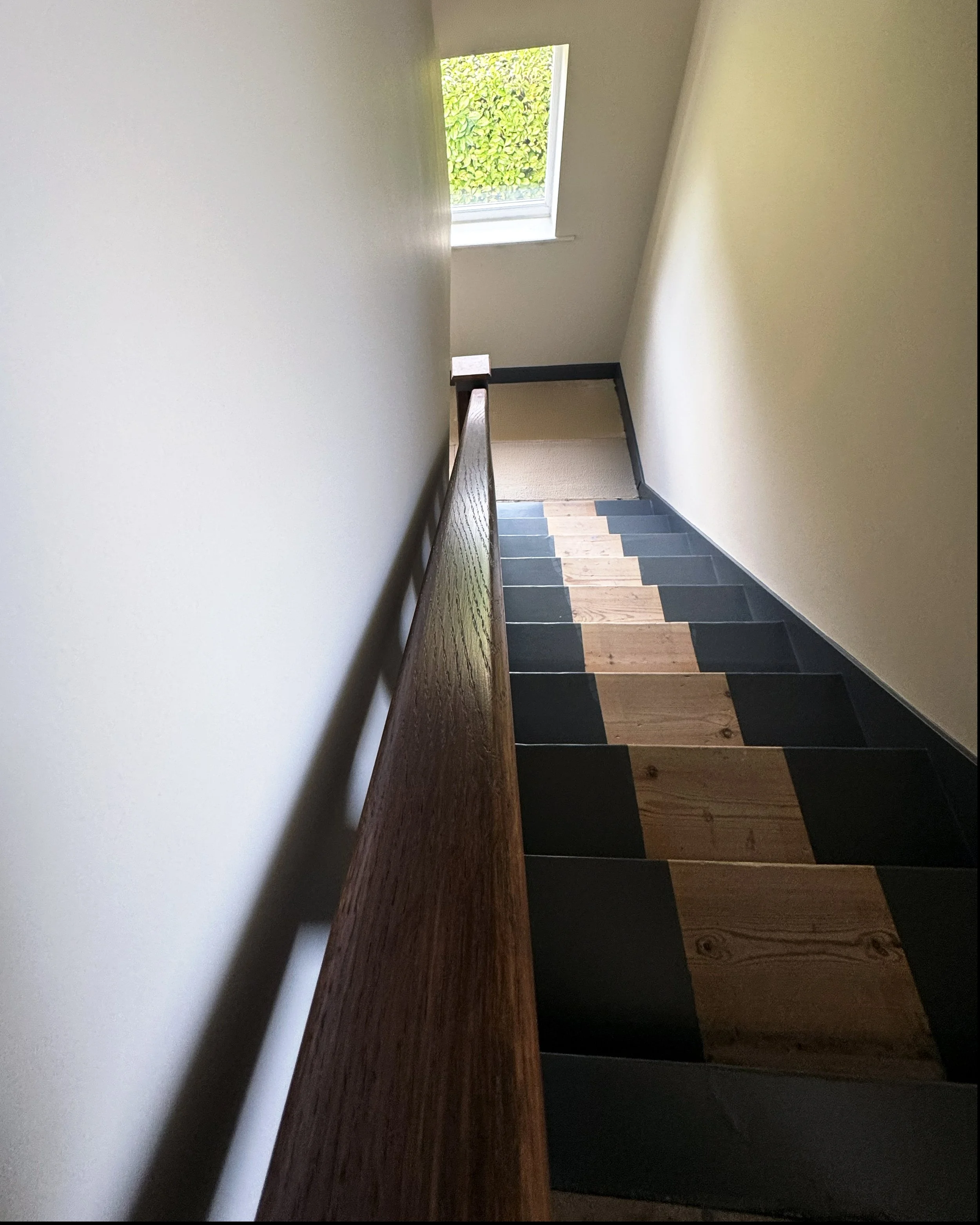 View of stairwell from above showing wooden handrail, black and natural wood checkered flooring, and window letting in natural light.