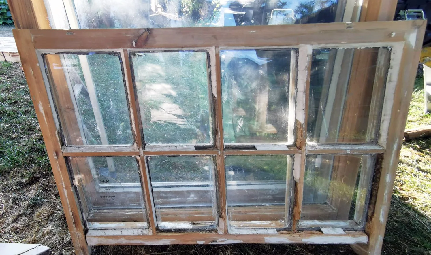 An old wooden window frame with multiple glass panes, some of which are cracked or broken, leaning against a wooden structure outdoors.
