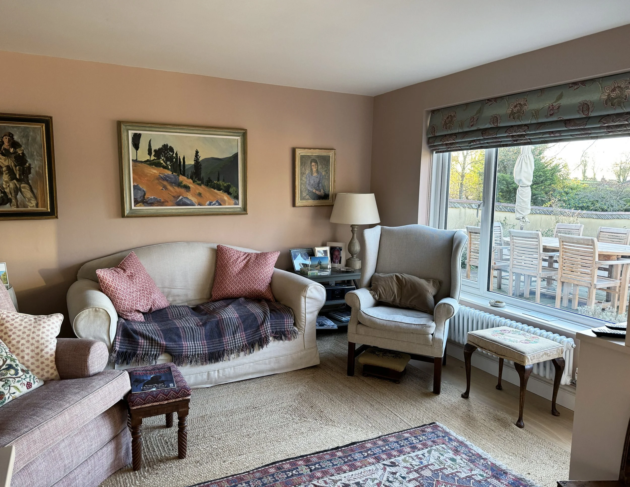 Living room with beige sofa and armchair, pink pillows, a blanket, framed artwork on the wall, a side table with family photos and a lamp, large window with patio outdoor furniture, and a rug on wooden floor.