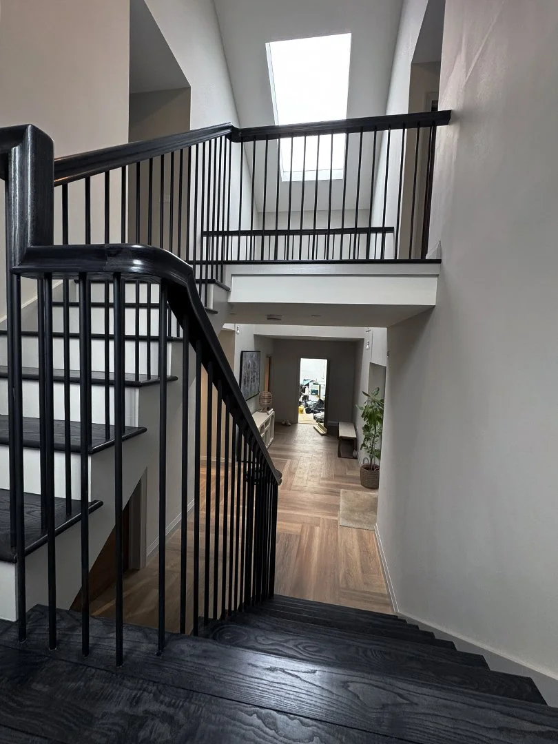 Interior view of a multi-level home with a staircase and black metal railings, leading to the ground floor with hardwood flooring, a hallway with furniture and decor, and skylights providing natural light.