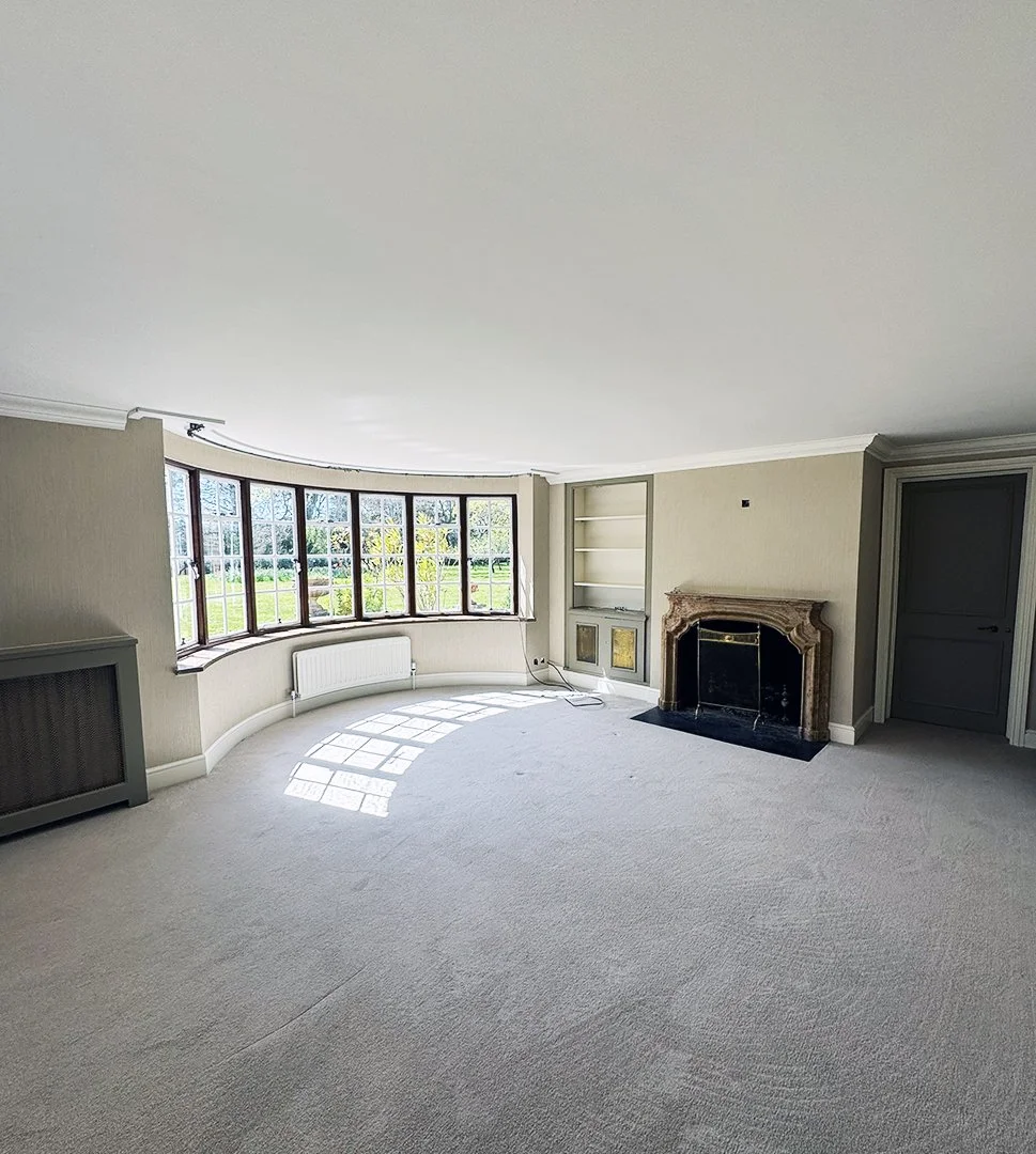 Empty living room with large bay window, beige walls, white carpet, a fireplace with an ornate marble mantel, built-in shelves, and a door to the right.