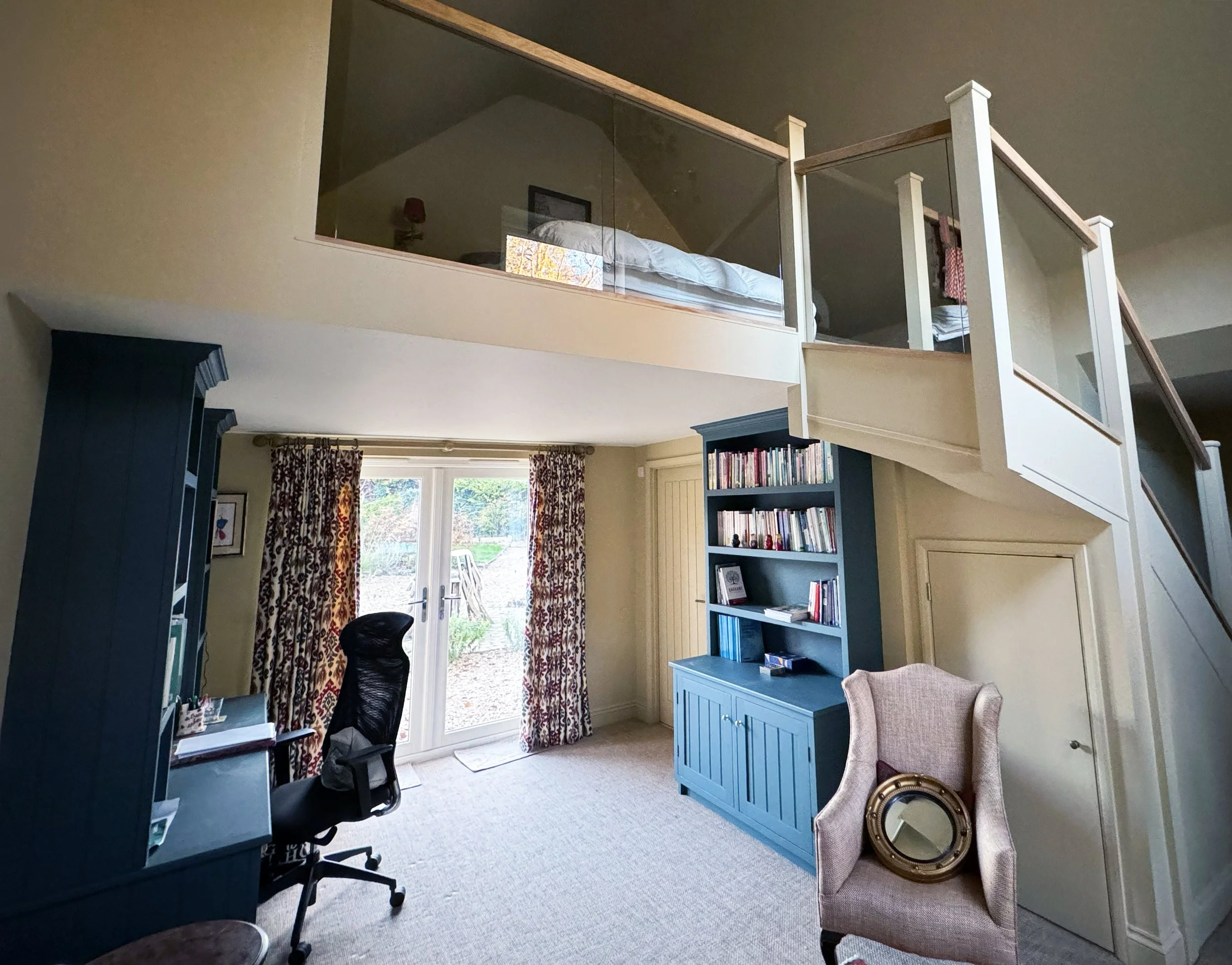 Room with blue bookshelf, beige armchair with mirror, black office chair, and glass doors leading outside, with an upper loft area visible.