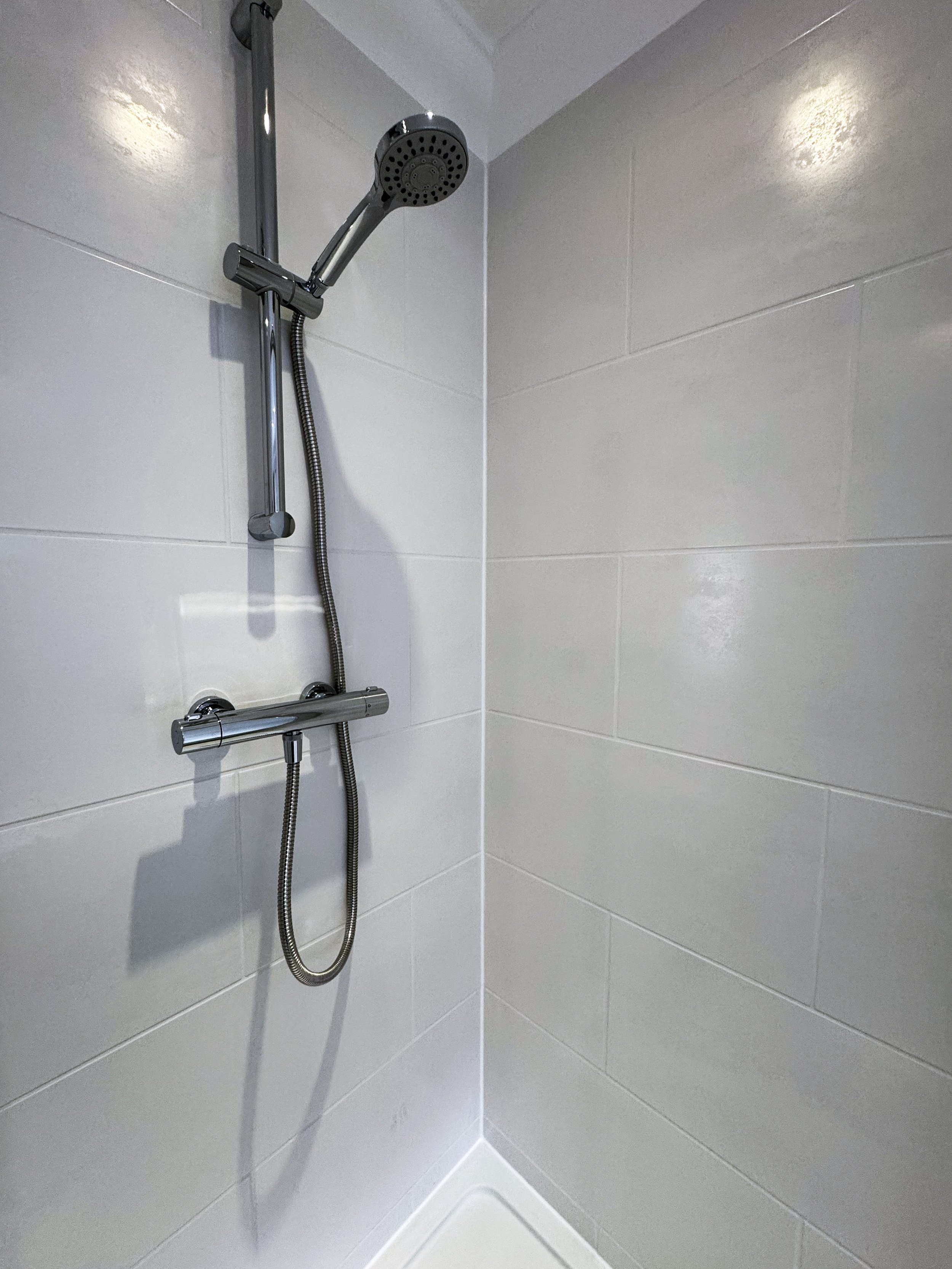 Empty shower with white tiled walls and a mounted chrome showerhead on a sliding rail, with a flexible hose and a wall-mounted handle.