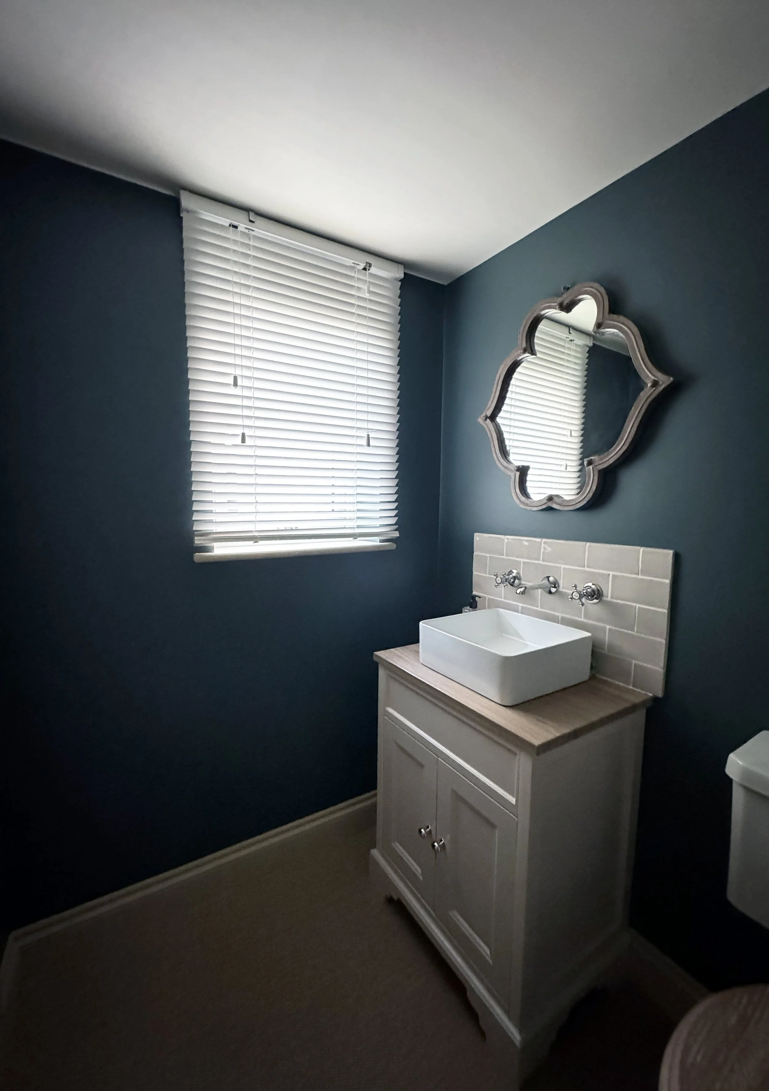 A small bathroom with dark blue walls, a window with white blinds, a white vanity with a vessel sink, a decorative mirror, and a beige tile backsplash.