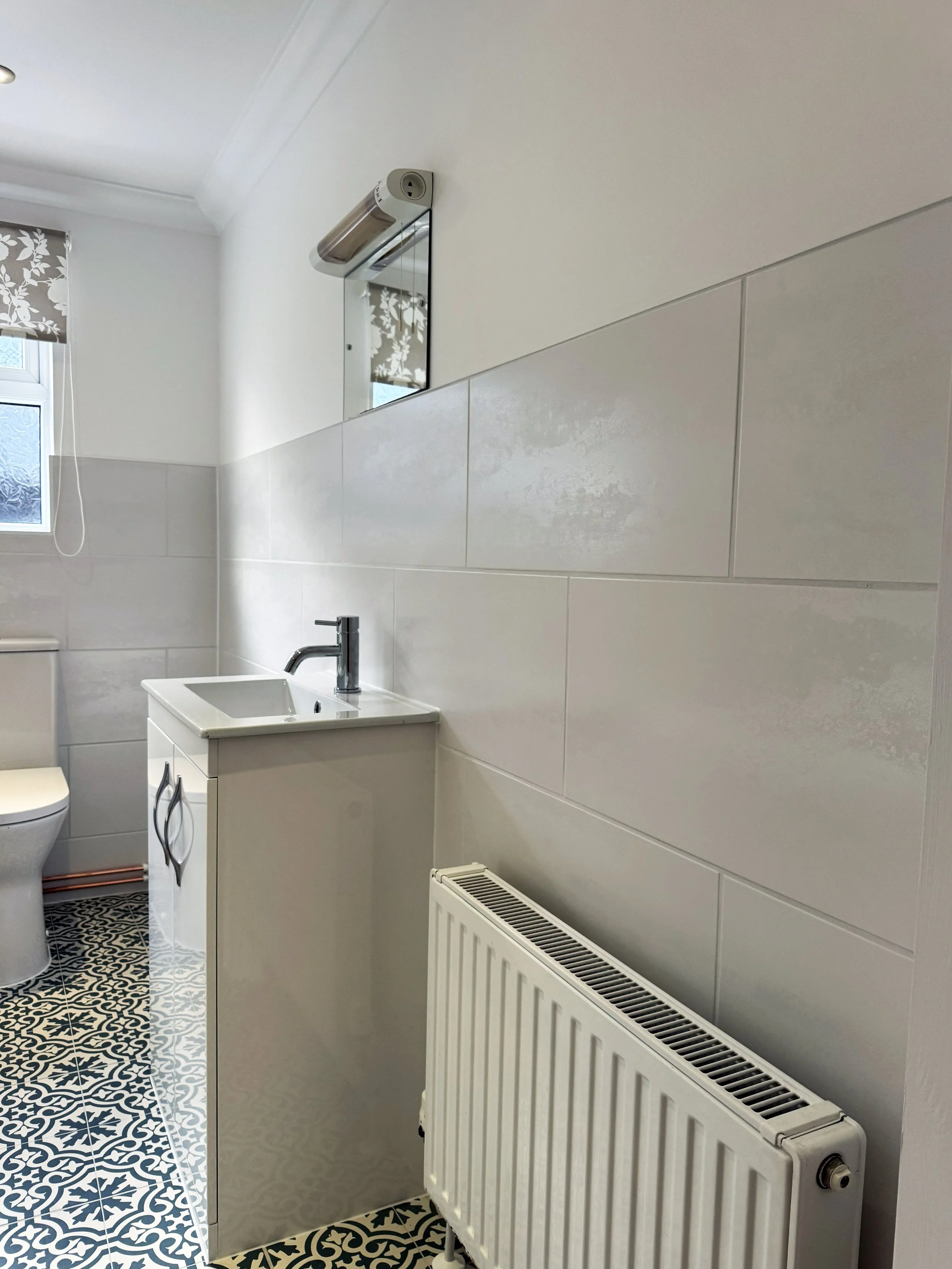 A small bathroom with a white sink cabinet, a wall-mounted mirror, a small window with floral curtains, and a radiator on the wall.