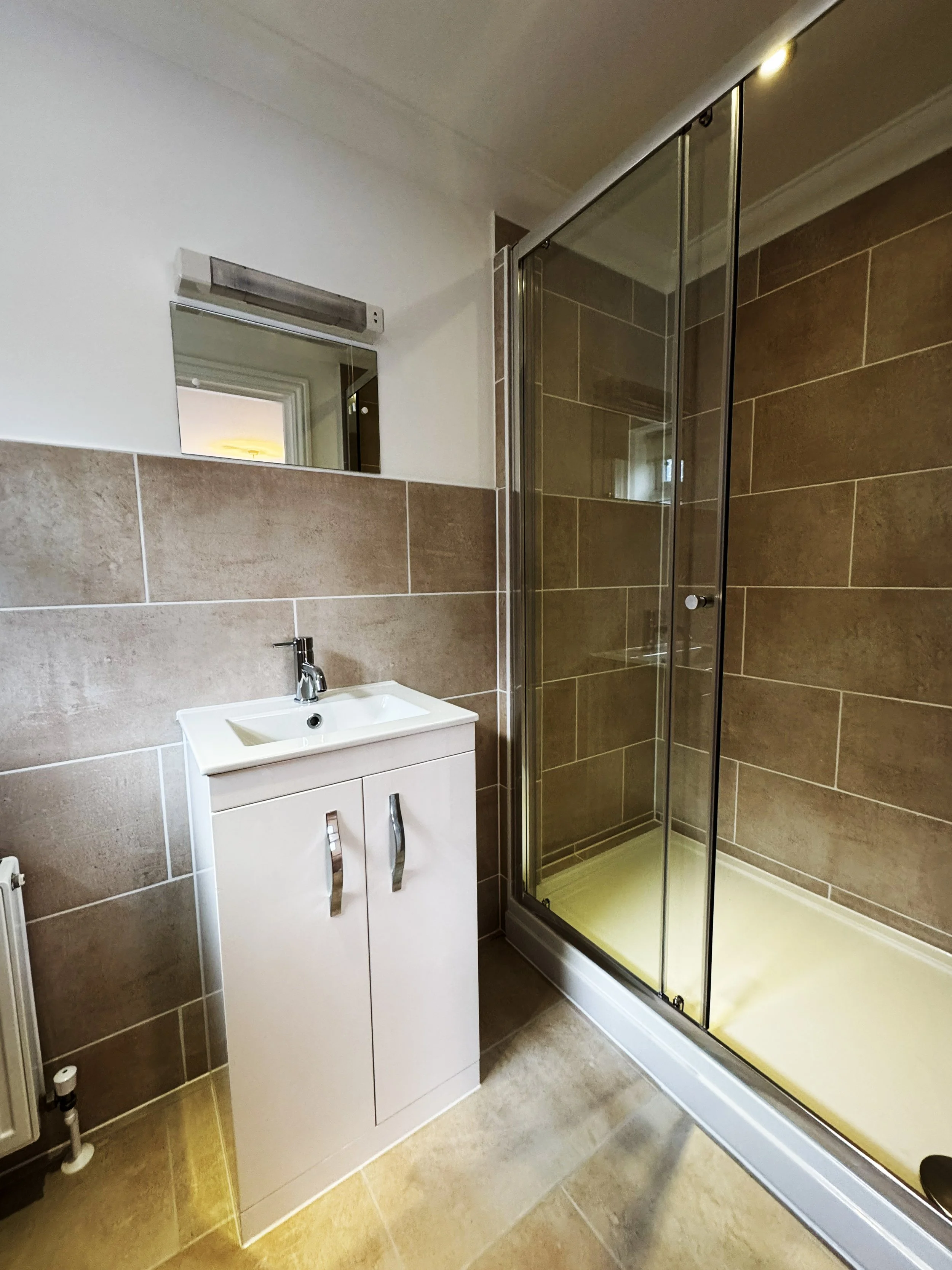 A bathroom with a small white sink cabinet, a mirror above it, and a glass-enclosed shower with brown tiled walls.
