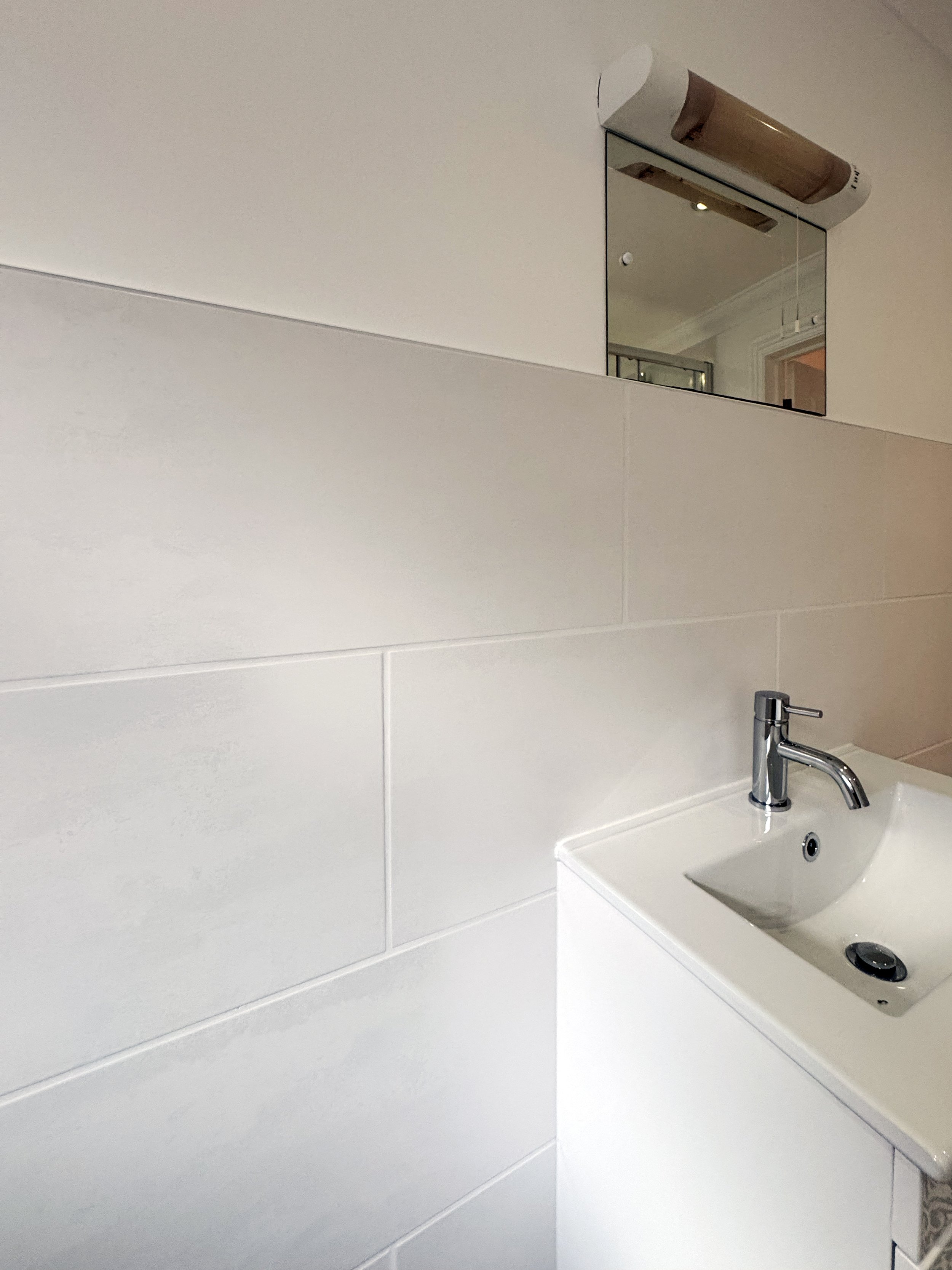 A small white bathroom sink with a modern chrome faucet and a circular drain, set against a light-colored tiled wall, with a mirror above and a paper towel dispenser on the wall.