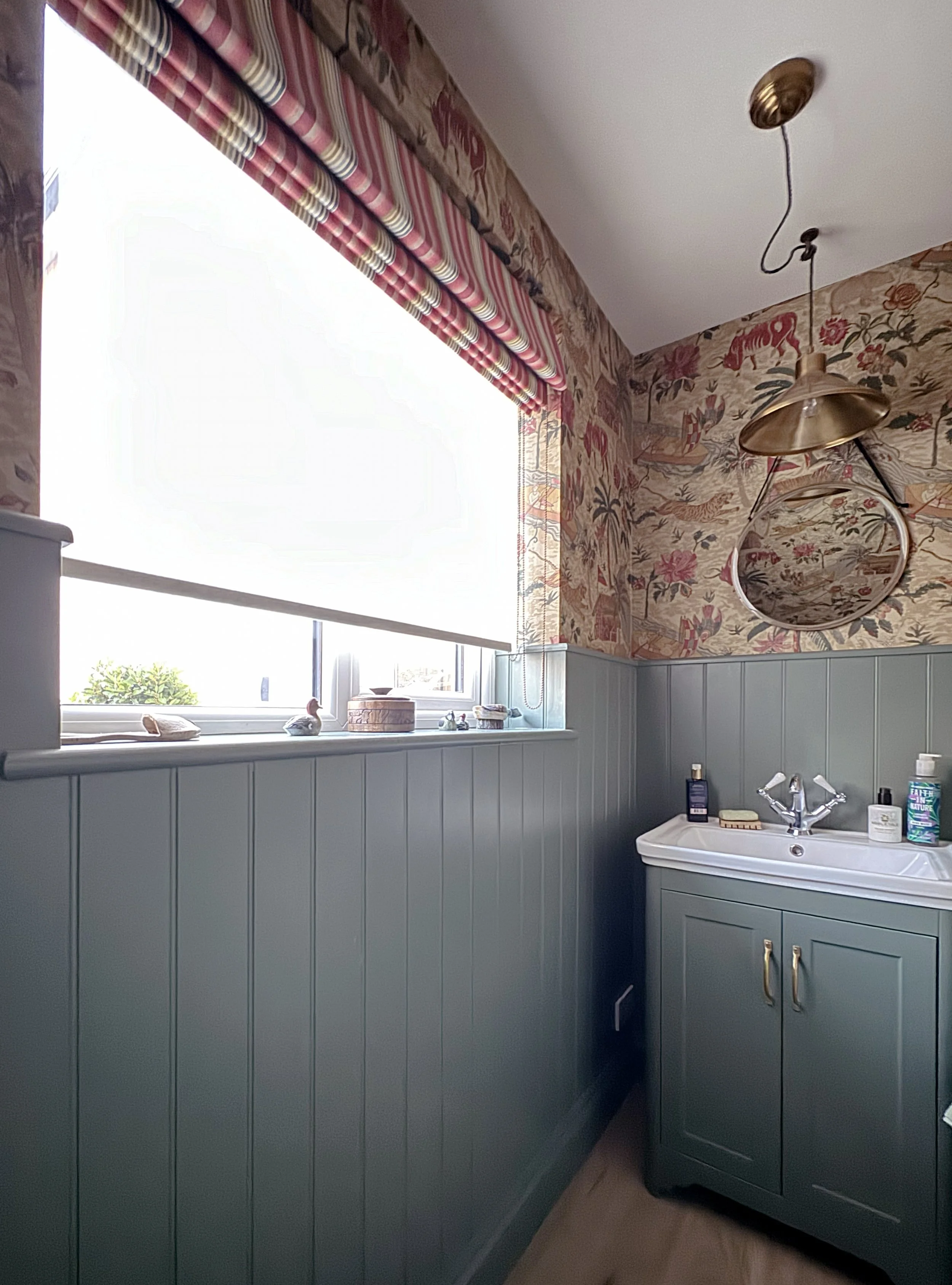 A bathroom corner with a window, a pale green cabinet with a white sink, and a patterned vintage wallpaper with elephants, lions, and acacia trees.
