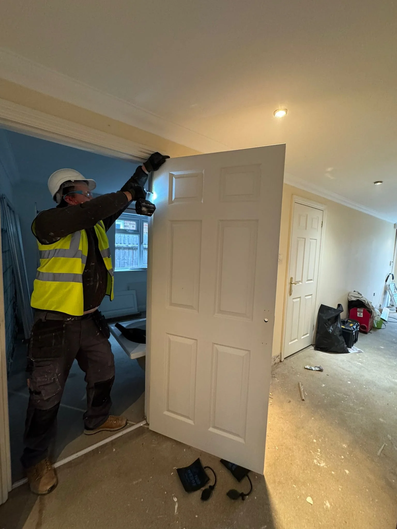 A construction worker in a yellow safety vest, helmet, and gloves installing or inspecting a household door with a power tool inside a house under renovation.