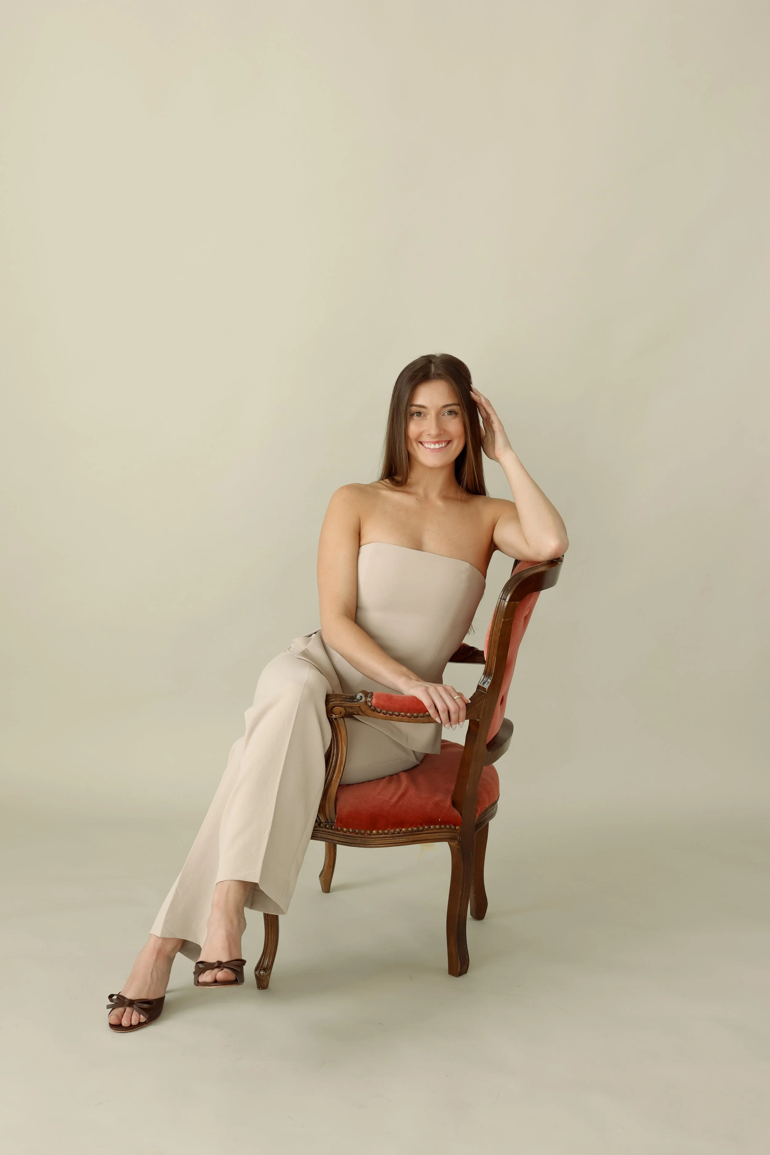 A woman sitting on a vintage wooden chair with red upholstery, smiling, with one hand touching her hair, wearing a strapless beige outfit and brown high heels.