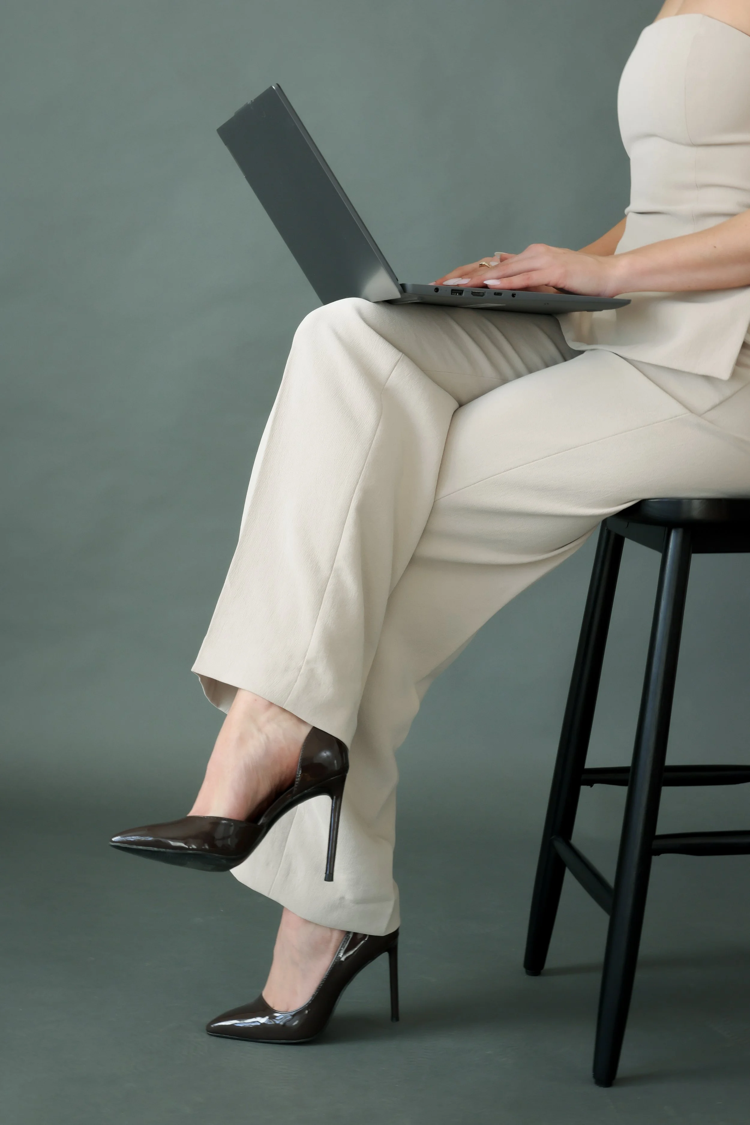 A woman sitting on a black barstool, wearing beige pants and a beige top, working on a laptop. She is wearing black high heels.
