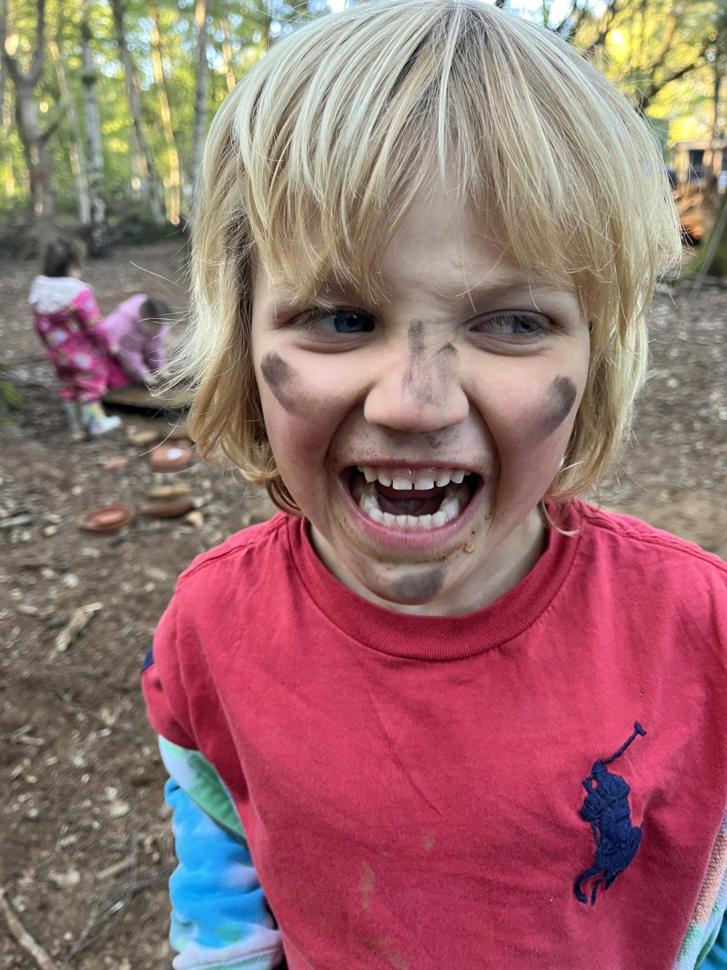 Close-up of a young blonde boy with paint smudges on his face, smiling widely with his mouth open, outdoors in a wooded area.