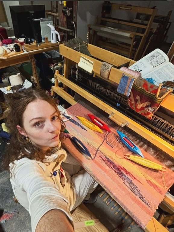 Ainsley taking a selfie, sitting infront of the loom with an active weaving visible