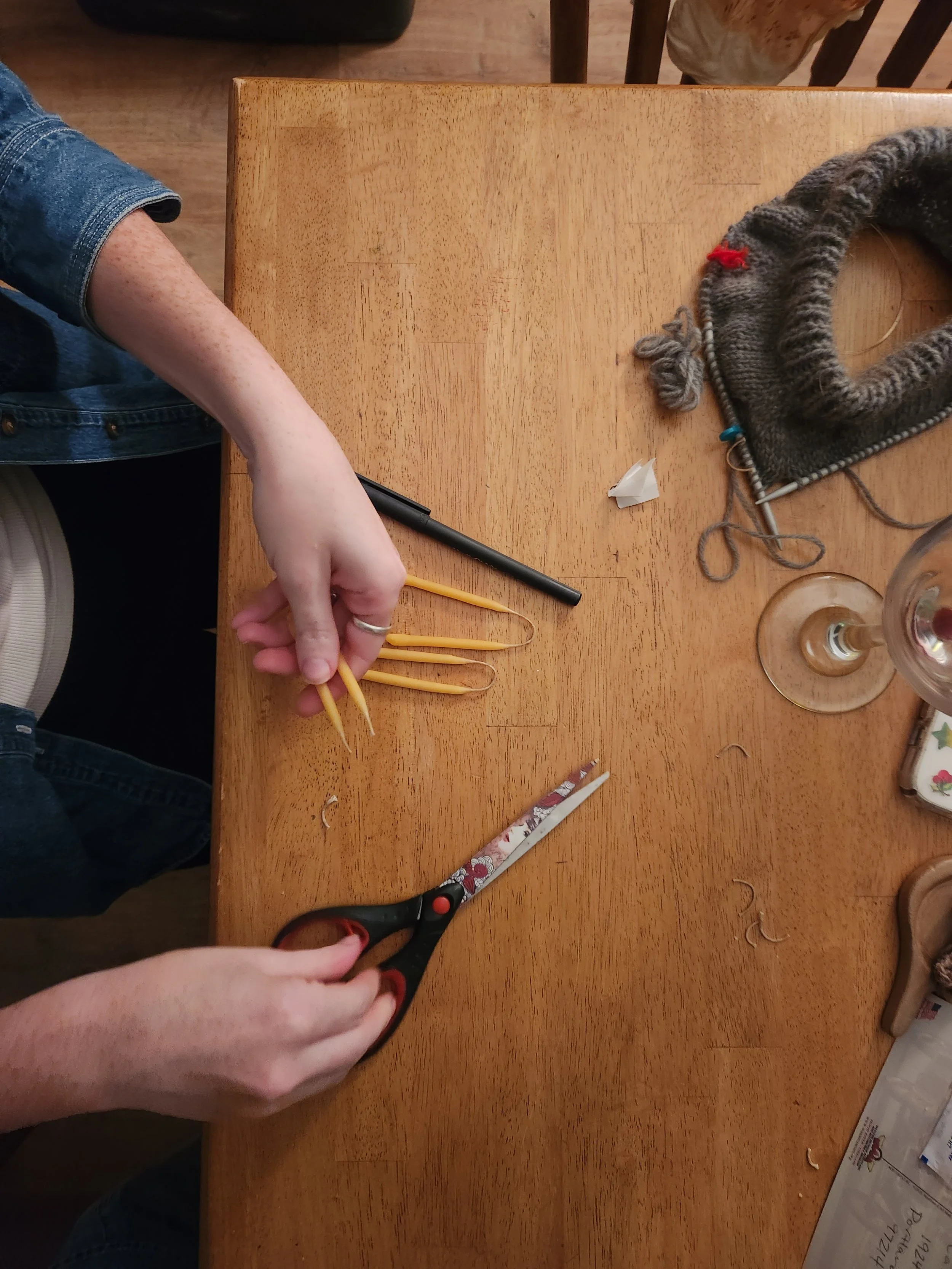 an aerial view of a someone using scissors to cut the wicks of hand-dipped birthday candles. in a corner, an in-progress knitting project and glass of wine are visible