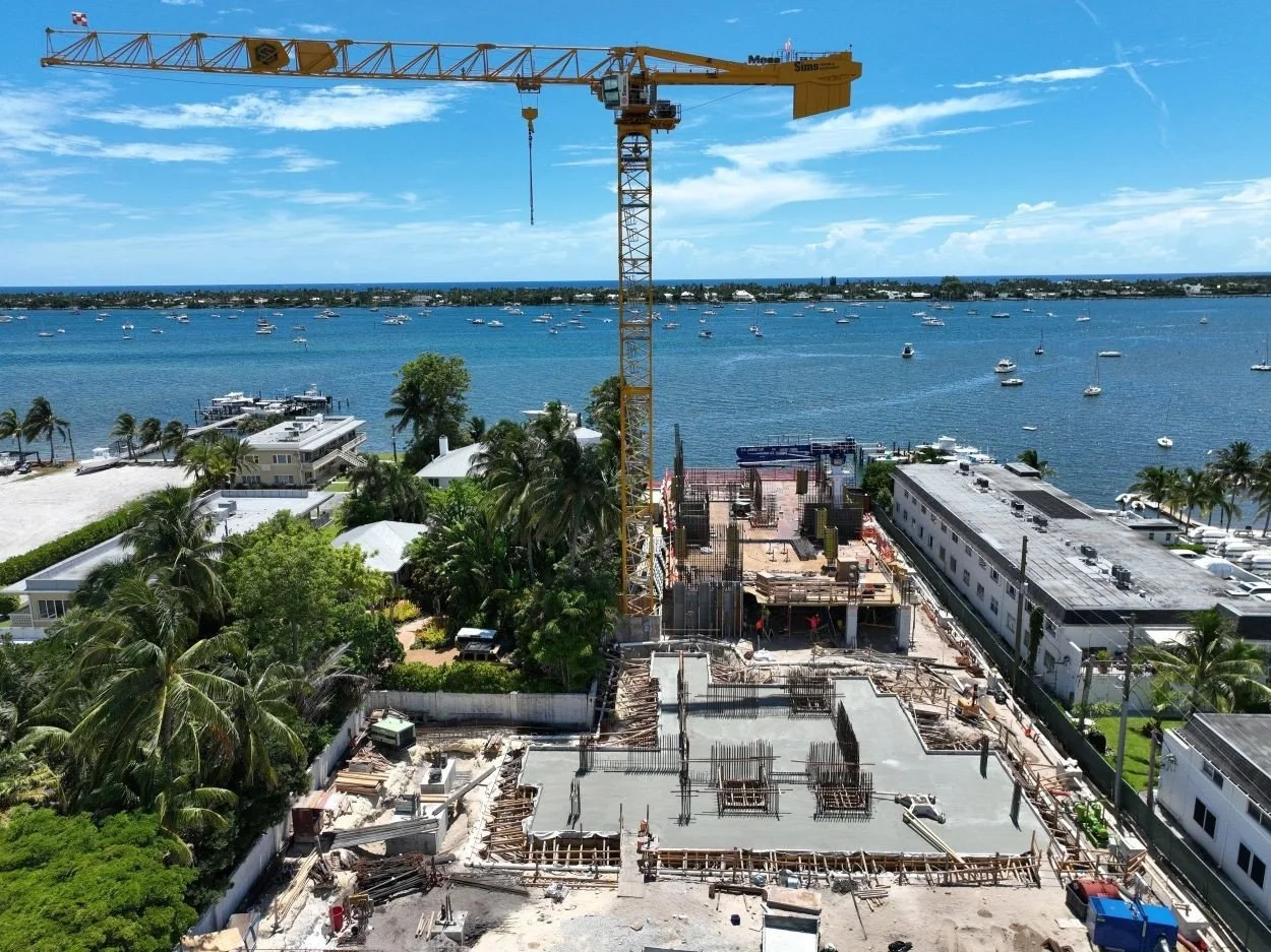 A construction crane works near the water, surrounded by construction materials and equipment on the site.