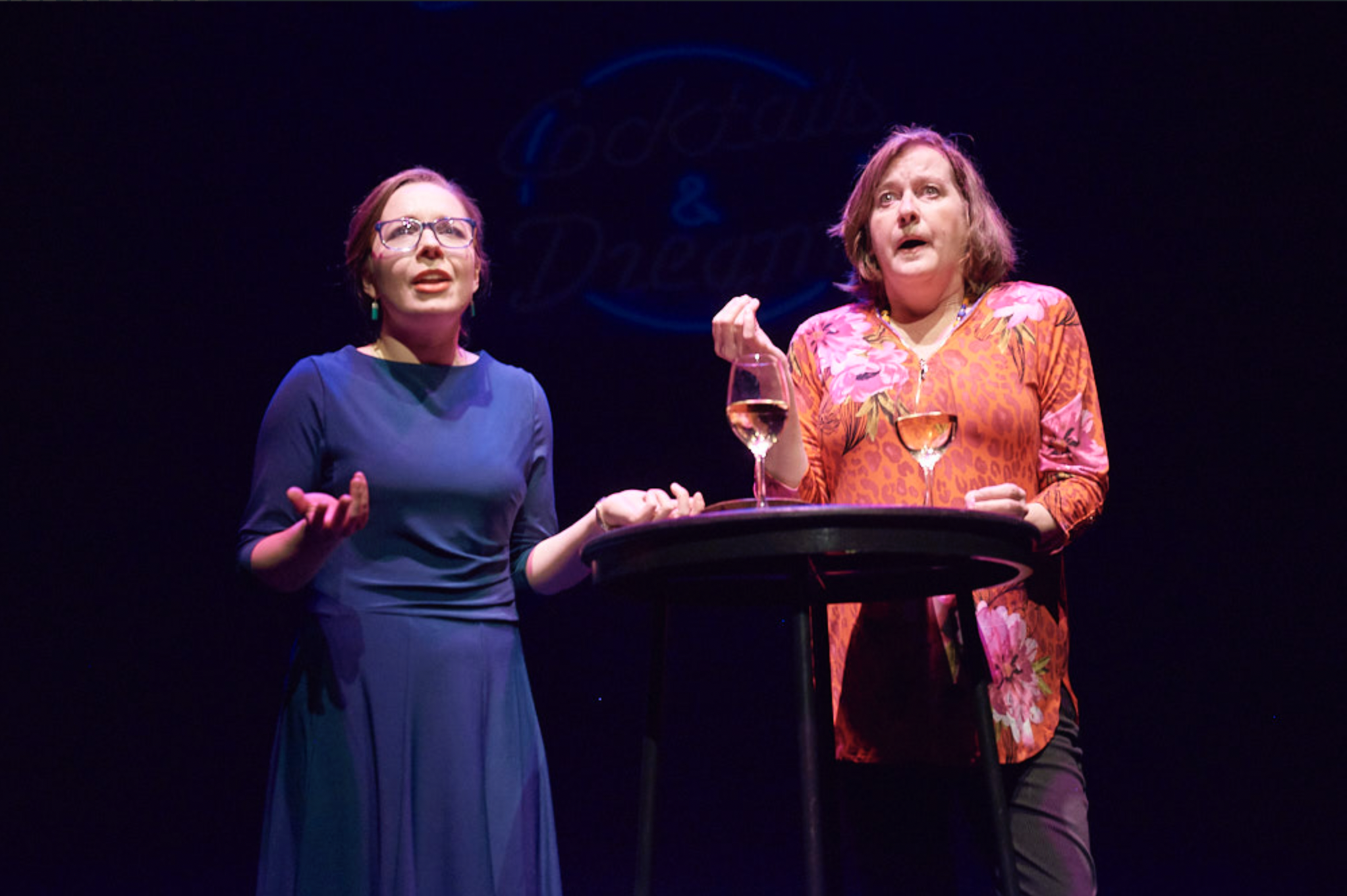 Two women stand on a stage with a table holding two glasses of white wine. One woman, on the left, wears a blue dress and glasses, while the woman on the right wears a colorful floral blouse. They appear to be engaged in a theatrical performance, wit