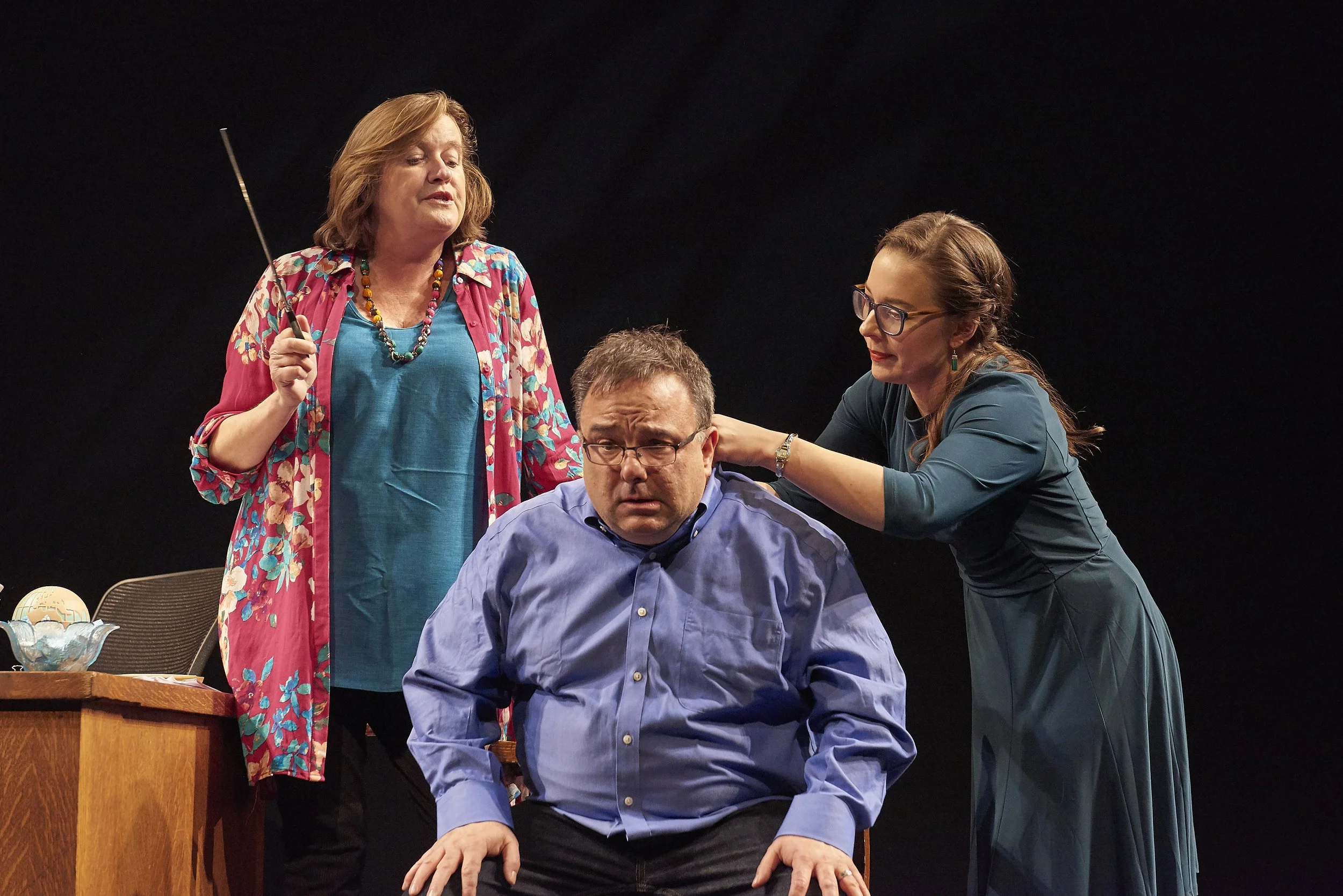 Three women and one man in a theatrical scene. The man is seated with a worried expression, while one woman touches his shoulder and another woman stands nearby with a wand or pointer, looking concerned.