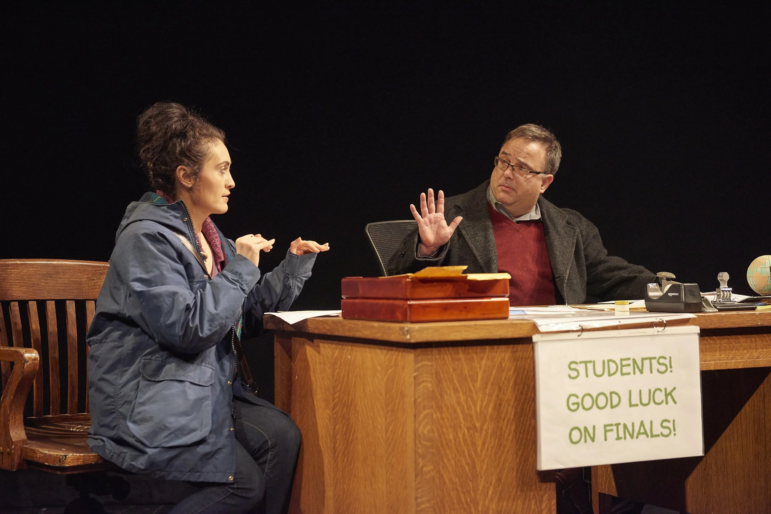 A young woman and an older man are engaged in a conversation in an office setting. The woman is gesturing with her hands, while the man is raising his hand in a stop or pause gesture. There are papers, boxes, and a globe on the desk. A sign on the de