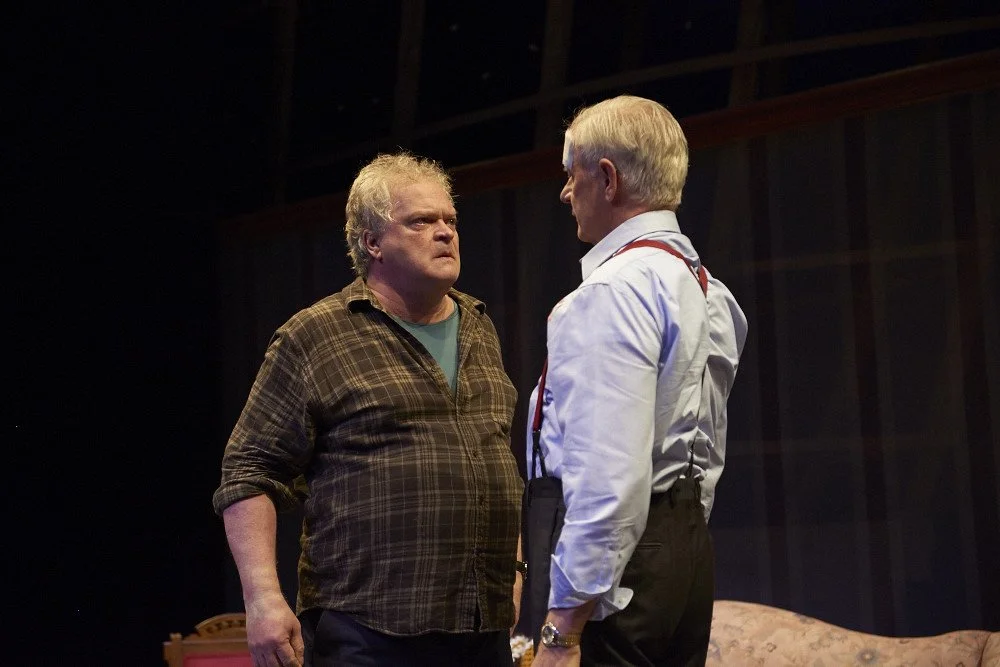 Two middle-aged men with serious expressions facing each other on a dimly lit stage.