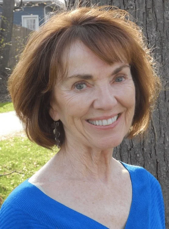 A smiling middle-aged woman with short auburn hair, wearing a blue top and earrings, standing outdoors near a tree with a house in the background.