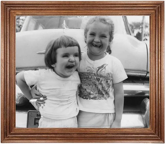 Two young girls smiling and laughing, standing in front of a vintage car, framed as a black and white photograph.