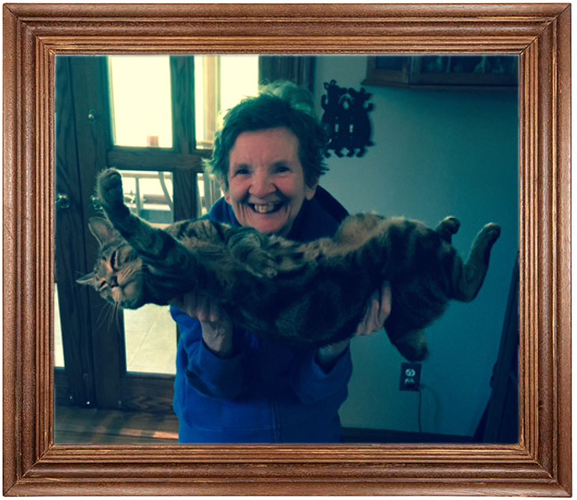 A woman holding a large, striped tabby cat horizontally in front of her, smiling indoors near a door and window.
