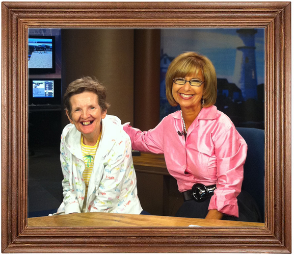 Two smiling women, one elderly with short gray hair wearing a floral top, and the other middle-aged woman with short light brown hair and glasses wearing a pink blouse, sitting at a table inside a room that appears to be a news studio or office.
