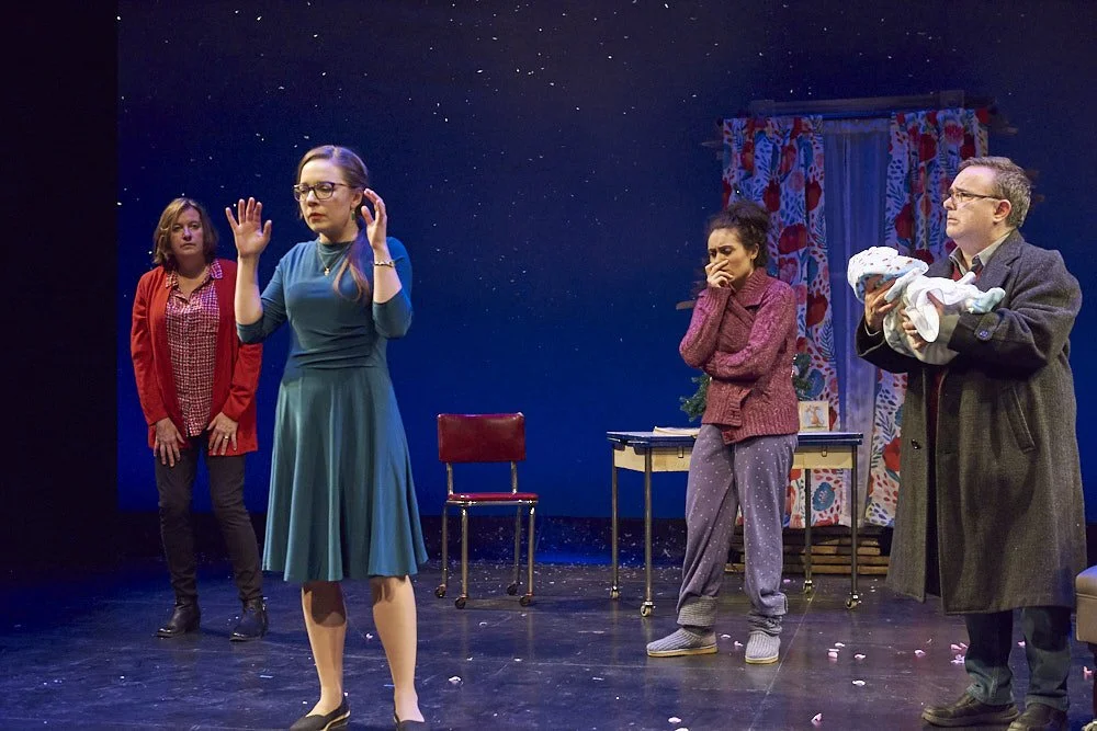Four actors on a stage during a theatrical performance. One woman in a blue dress appears to be speaking or acting, while the others watch. The background is a decorated set with a curtain and a table with small objects.