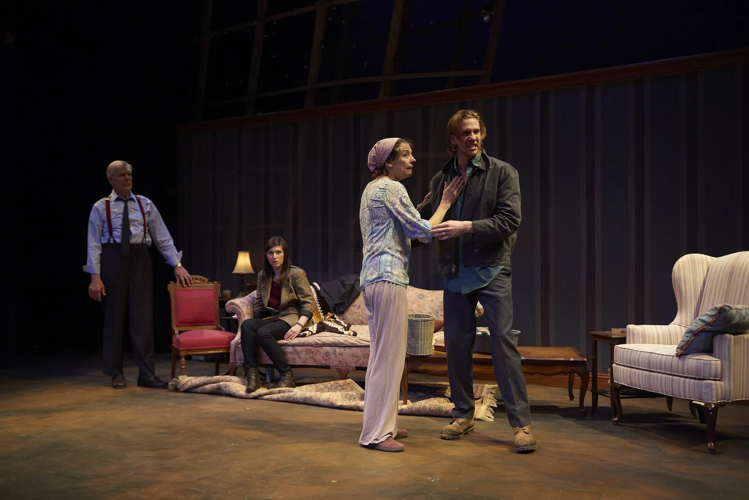 Four actors perform on a stage set like a living room. An older woman and a young man are in the foreground, holding each other. A young woman sits on a sofa behind them, while a man stands nearby, observing.