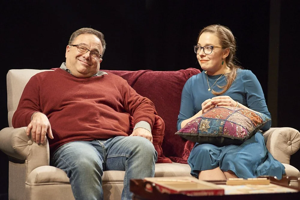 A man and woman sitting on a beige sofa with a red backrest in a dark room. The man is wearing glasses, a red sweater, and jeans, smiling at the camera. The woman is wearing glasses, a blue dress, and holding a colorful pillow, looking at the man wit