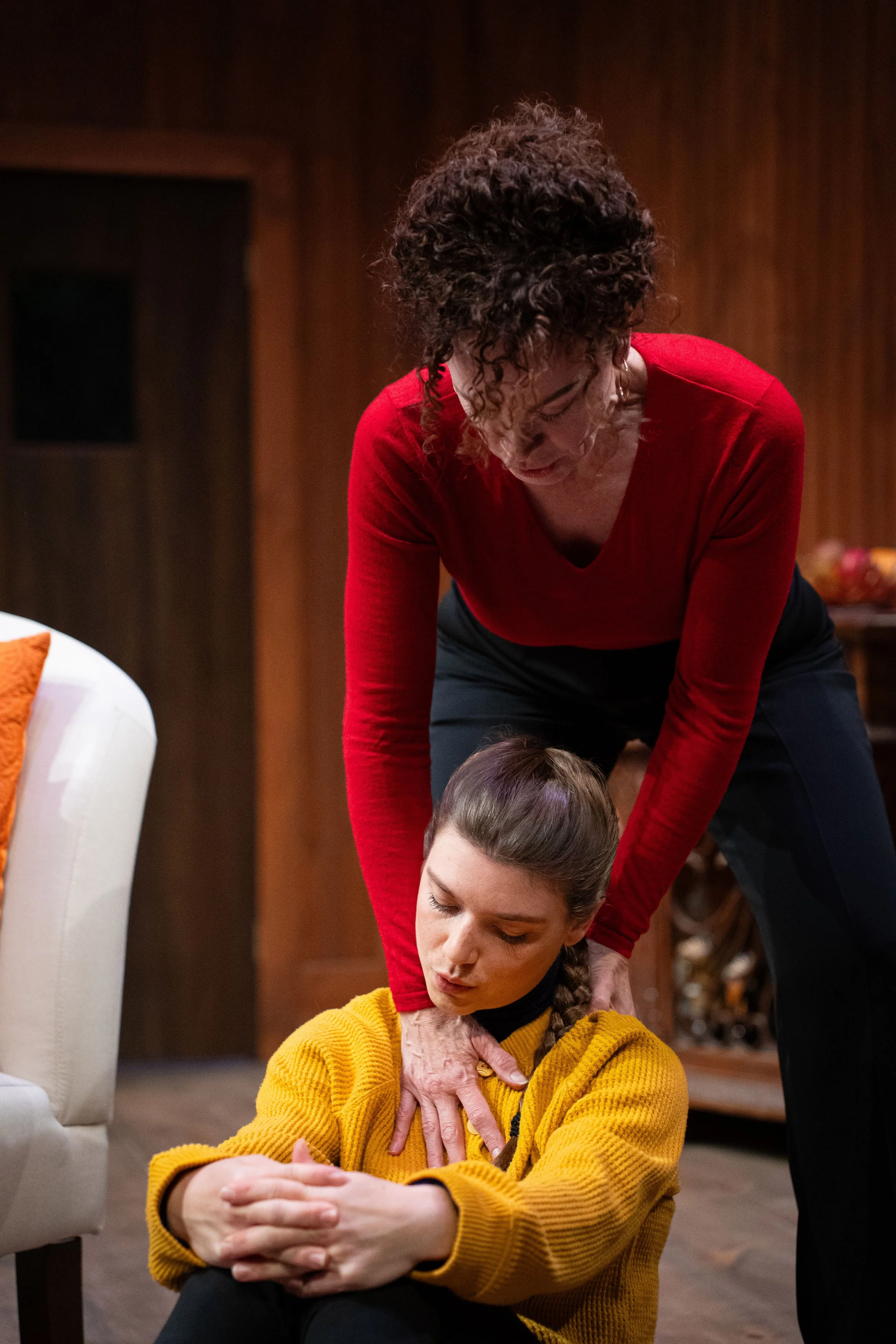 A woman in a red top is providing emergency first aid to a young woman in a yellow sweater who is sitting on the floor, appearing unresponsive. The scene suggests a medical emergency training or response.