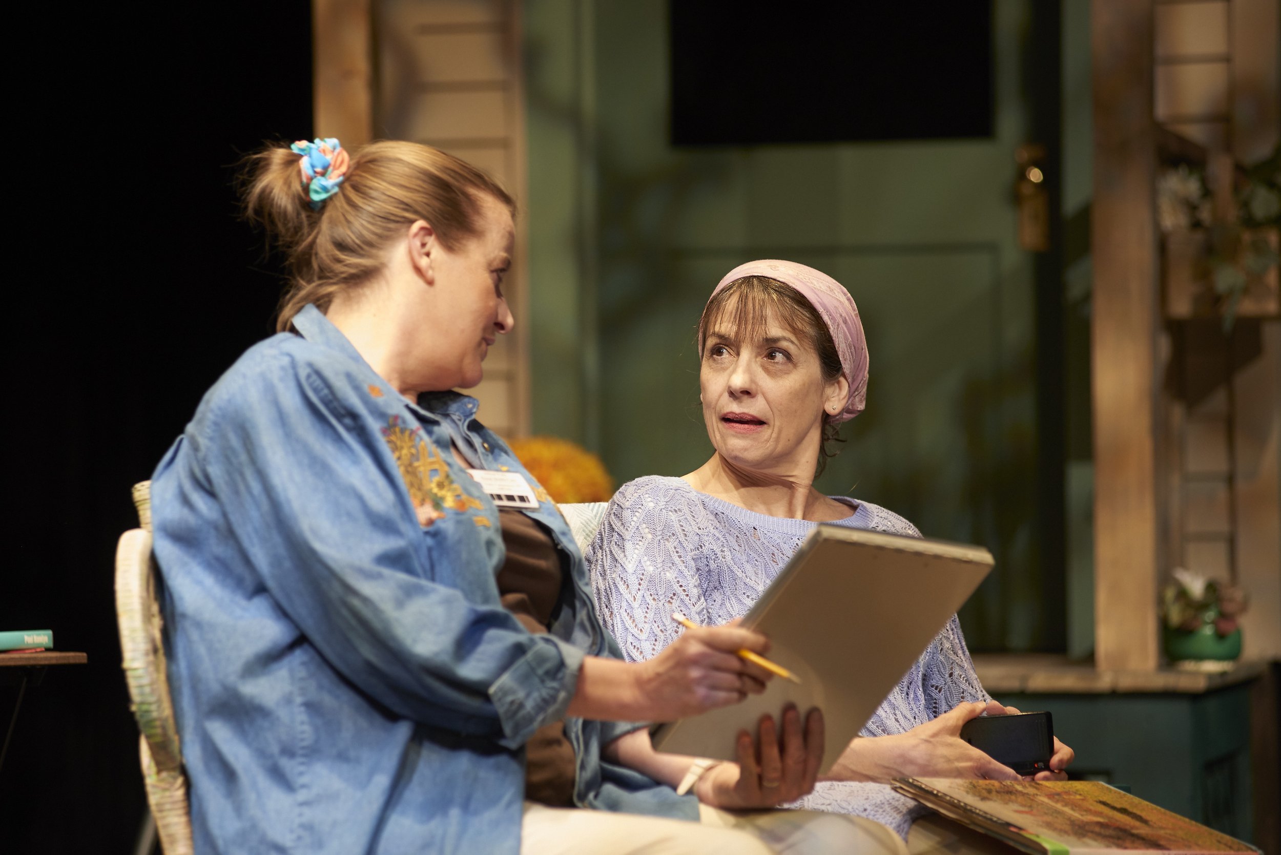 Two women sitting on a bench, engaged in conversation. The woman on the left has red hair tied with a colorful scrunchie and is wearing a denim jacket. The woman on the right has short hair, a pink headscarf, and is holding a notepad and a pen, with 