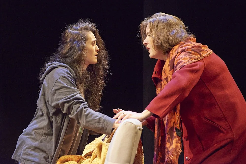 Two women engaged in a heated conversation, one leaning forward and holding the other's hands, on a stage with a black background.