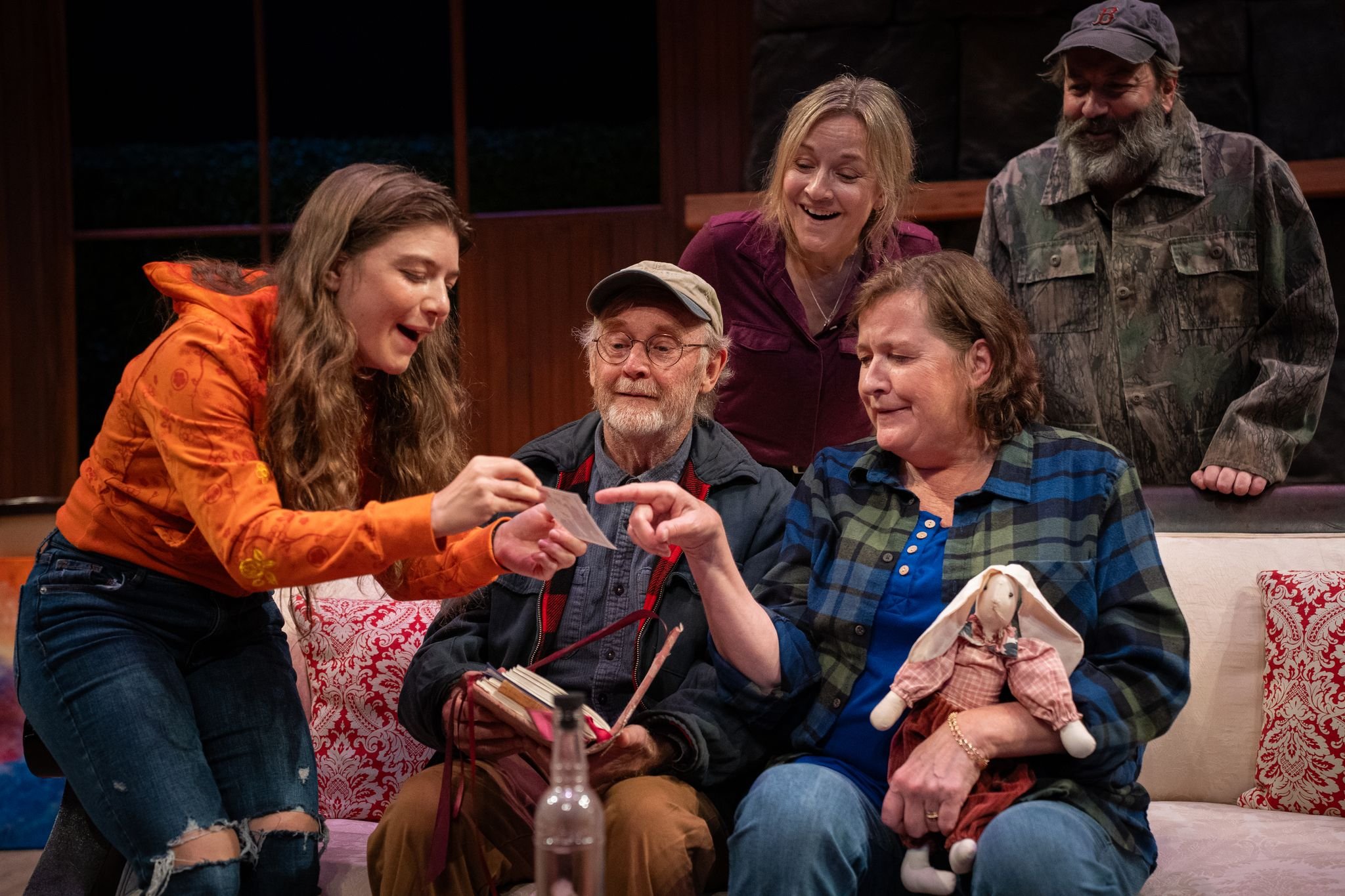 Group of five people sitting and standing on a couch, looking at a photo or document, smiling and pointing, in a cozy indoor setting. One woman holds a stuffed rabbit.