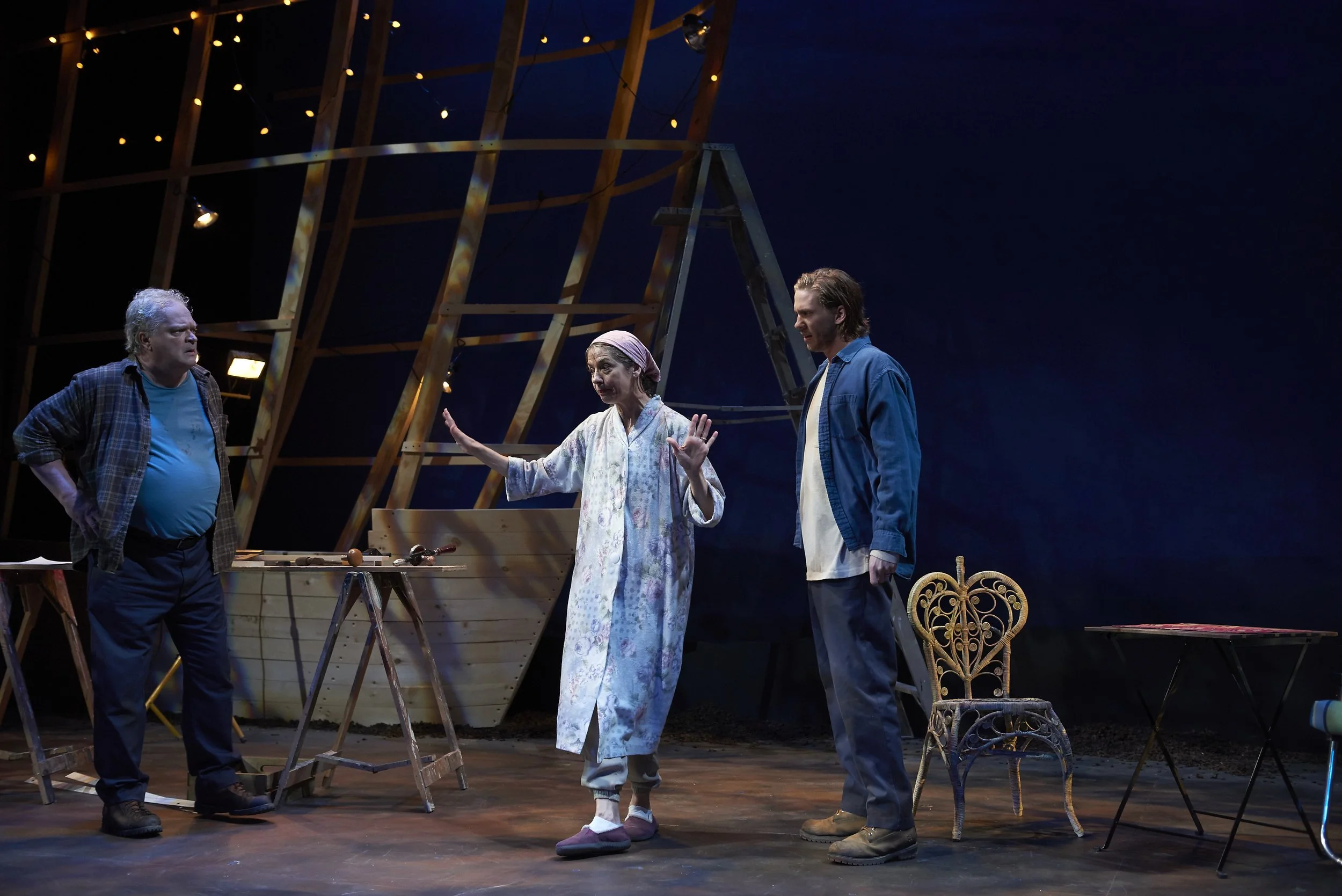 Three actors on stage during a theater performance, with construction materials and furniture around them, under stage lighting.