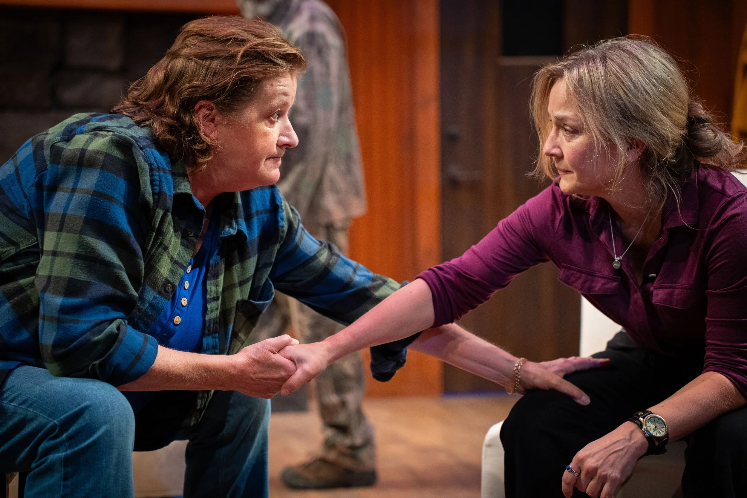 Two women are sitting on chairs, facing each other, holding hands, and making eye contact in a warm emotional moment, with a rustic wooden interior background.