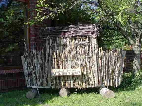 A small wooden structure with a bamboo fence around it, situated on wheels with logs underneath. A sign on the fence reads, 'Have you read the book?'