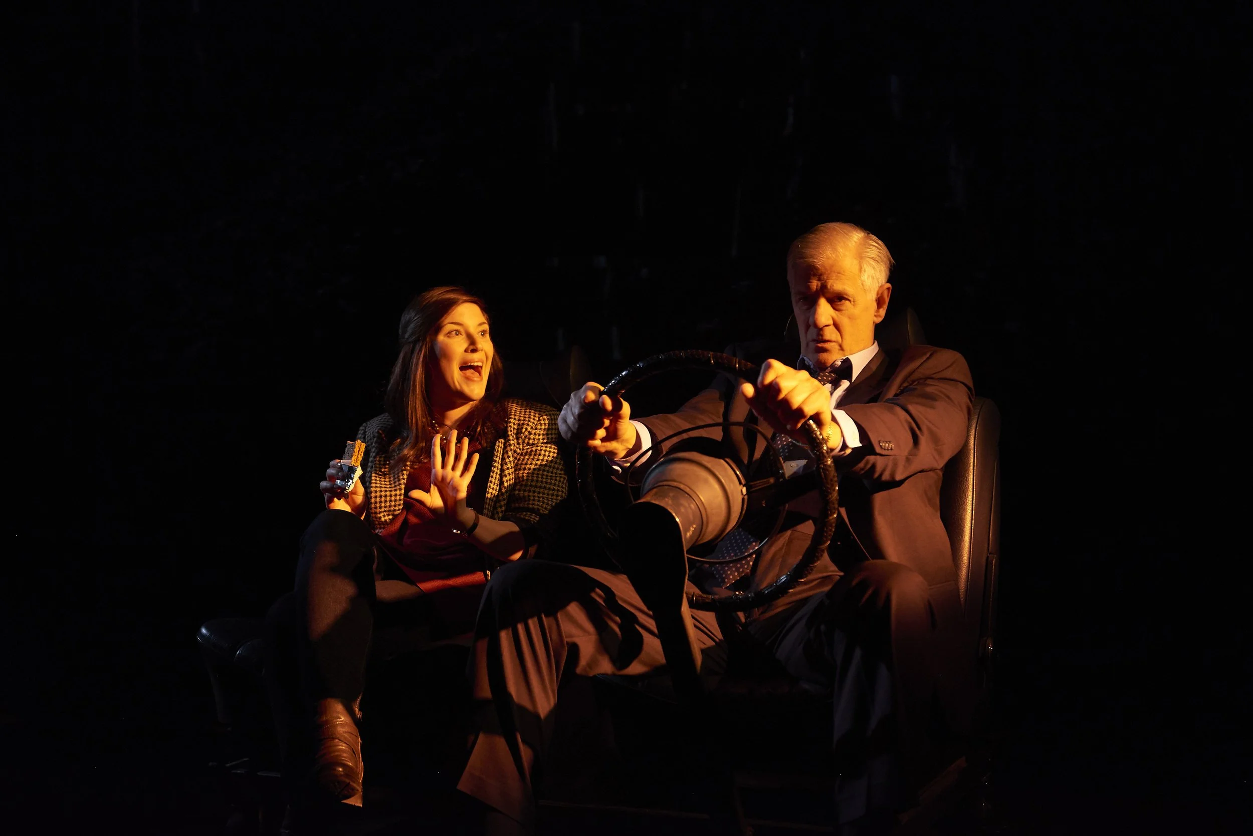 A woman with brown hair, wearing a checkered blazer, is sitting and talking animatedly, holding a small object in her right hand, while a man with gray hair, wearing a suit, is sitting with his hands on a steering wheel, looking serious. They are ill