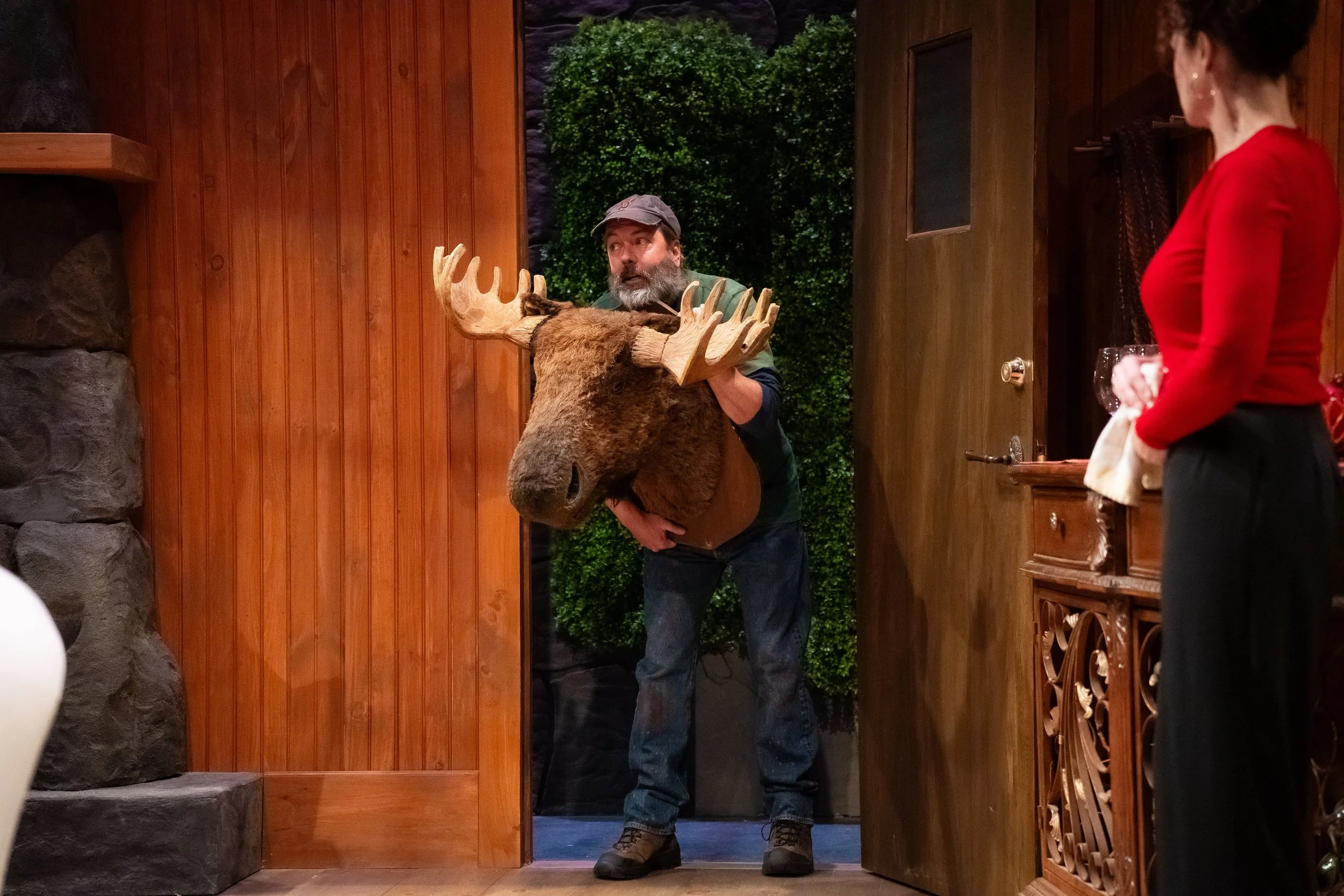 A man holding a moose head with large antlers on his shoulders, standing at a doorway in a cozy wooden interior, with a woman holding a glass of wine facing him.