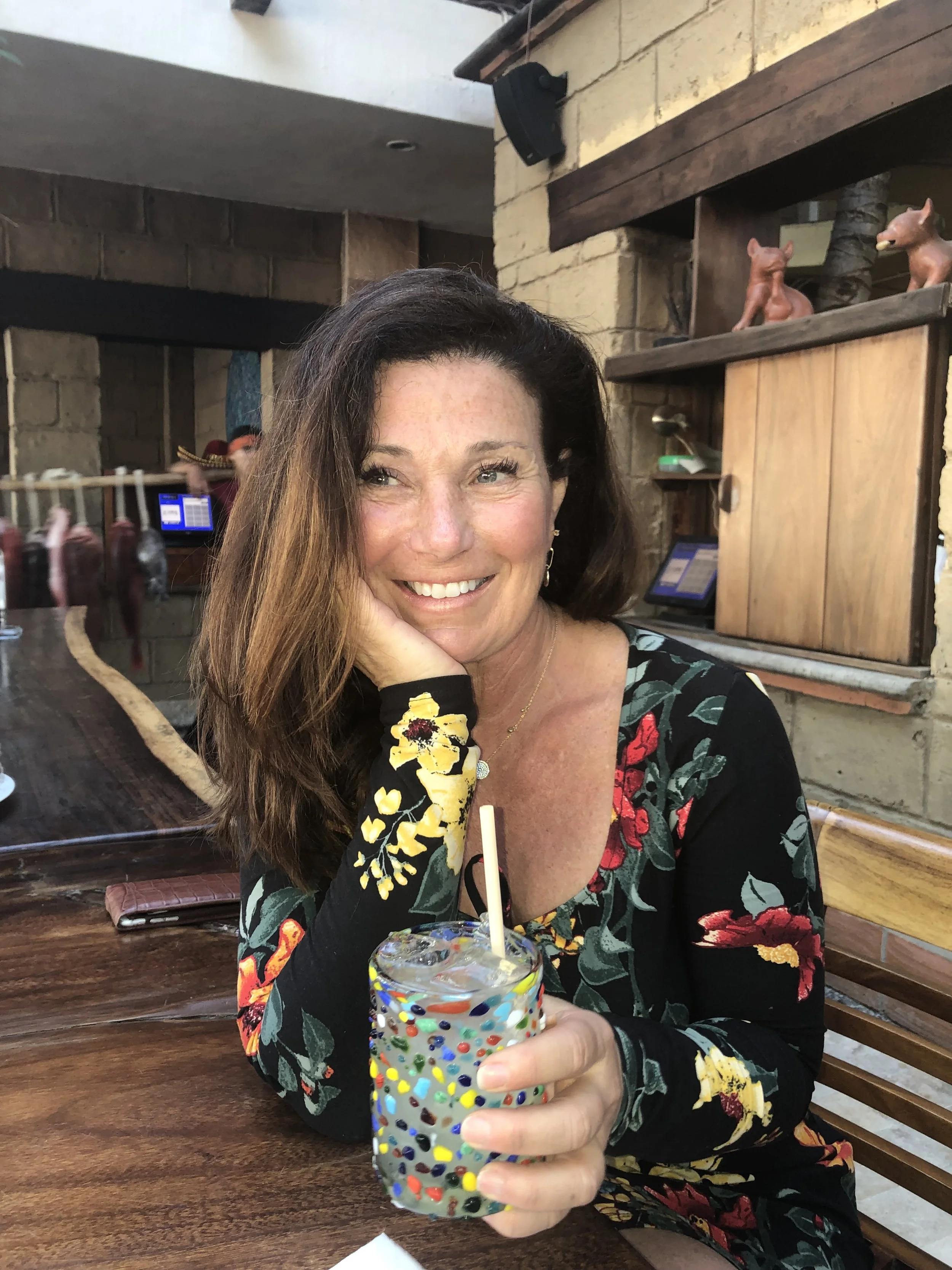 Chef Hana smiling at the camera, holding a glass of colorful sprinkle-topped drink, sitting at a wooden table in a cozy restaurant with a brick and wooden interior.
