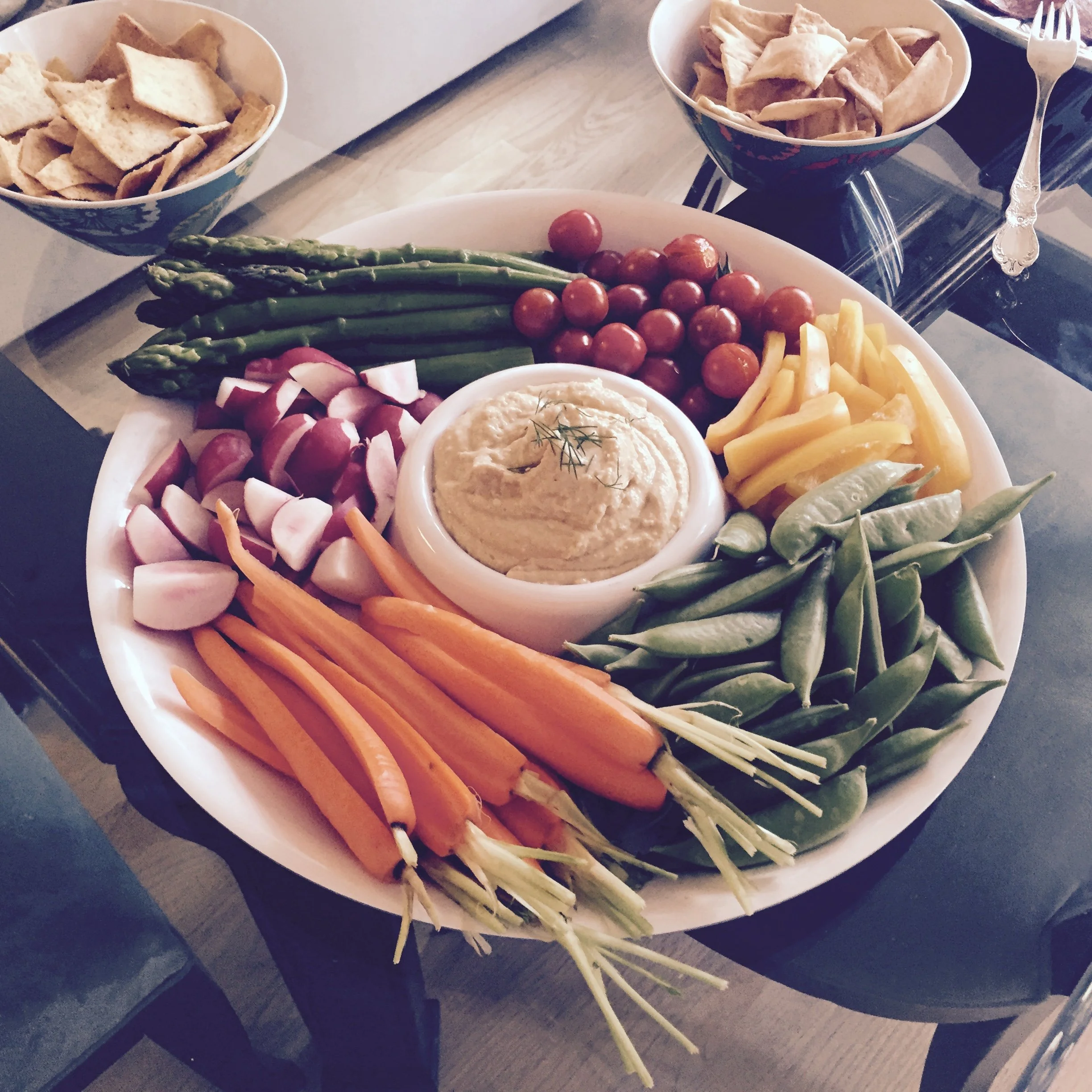 A large white platter of fresh vegetables including cherry tomatoes, asparagus, radishes, carrots, yellow bell peppers, and snap peas, with a bowl of hummus in the center. Two bowls of pita chips are seen in the background.