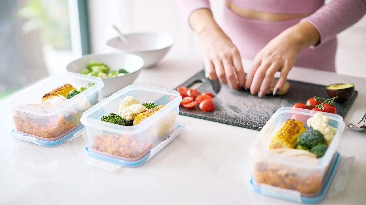Person preparing vegetables on a black cutting board with cherry tomatoes, avocado, and cauliflower, while pre-packed meal containers with broccoli, cauliflower, corn, and grilled chicken are on the white kitchen counter.
