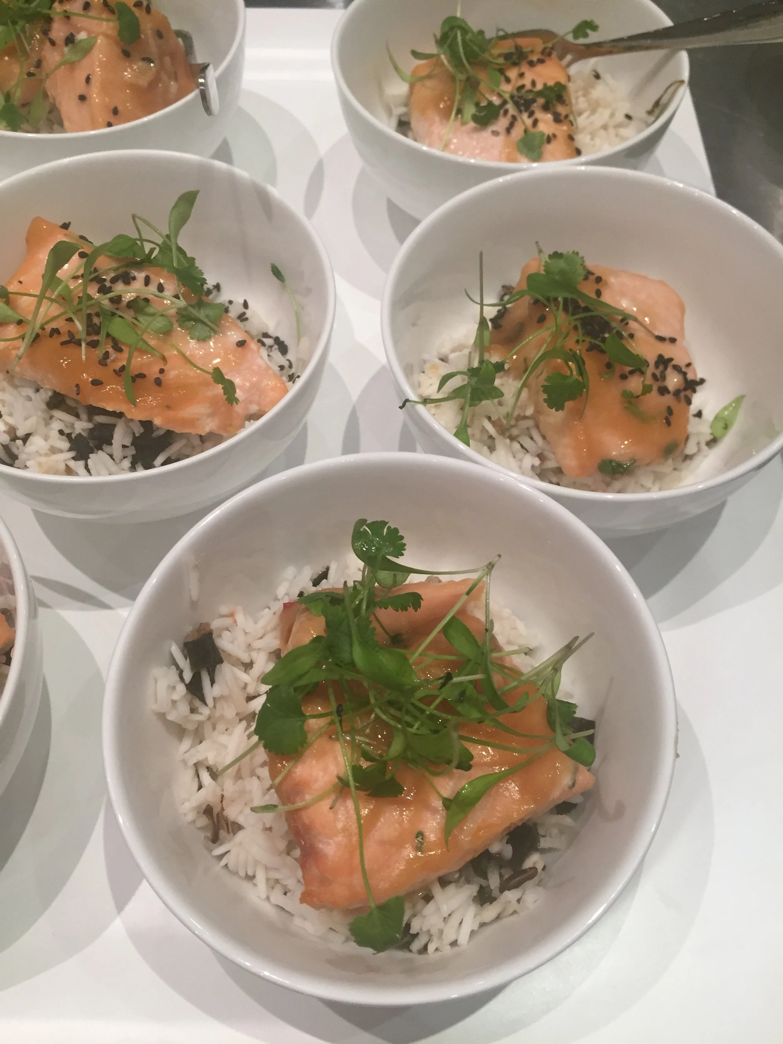 Bowls of rice topped with cooked salmon fillets, garnished with microgreens and black sesame seeds.