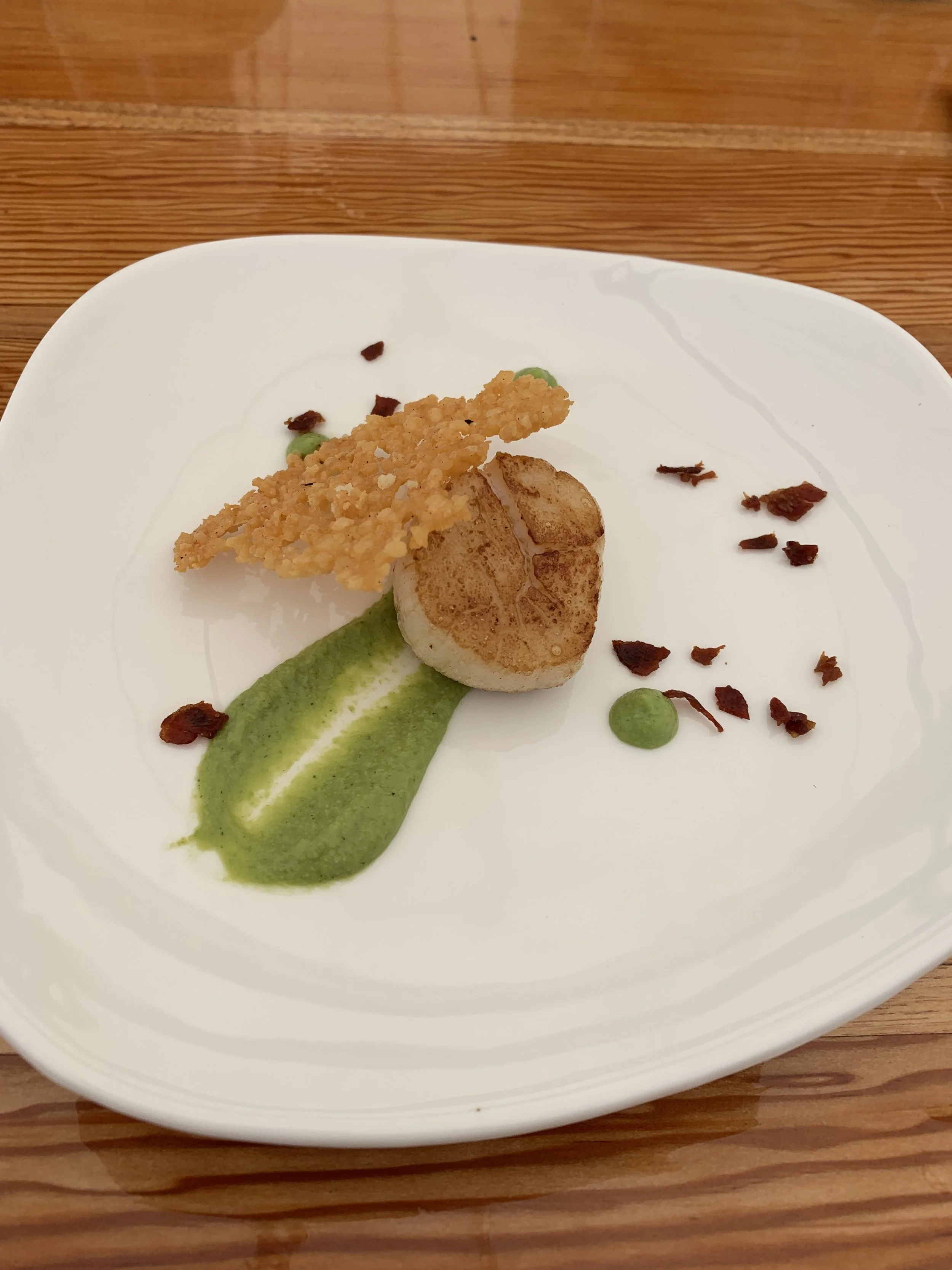 A plated gourmet dish with seared scallop, green sauce, crispy garnish, and small food bits on a white square plate.