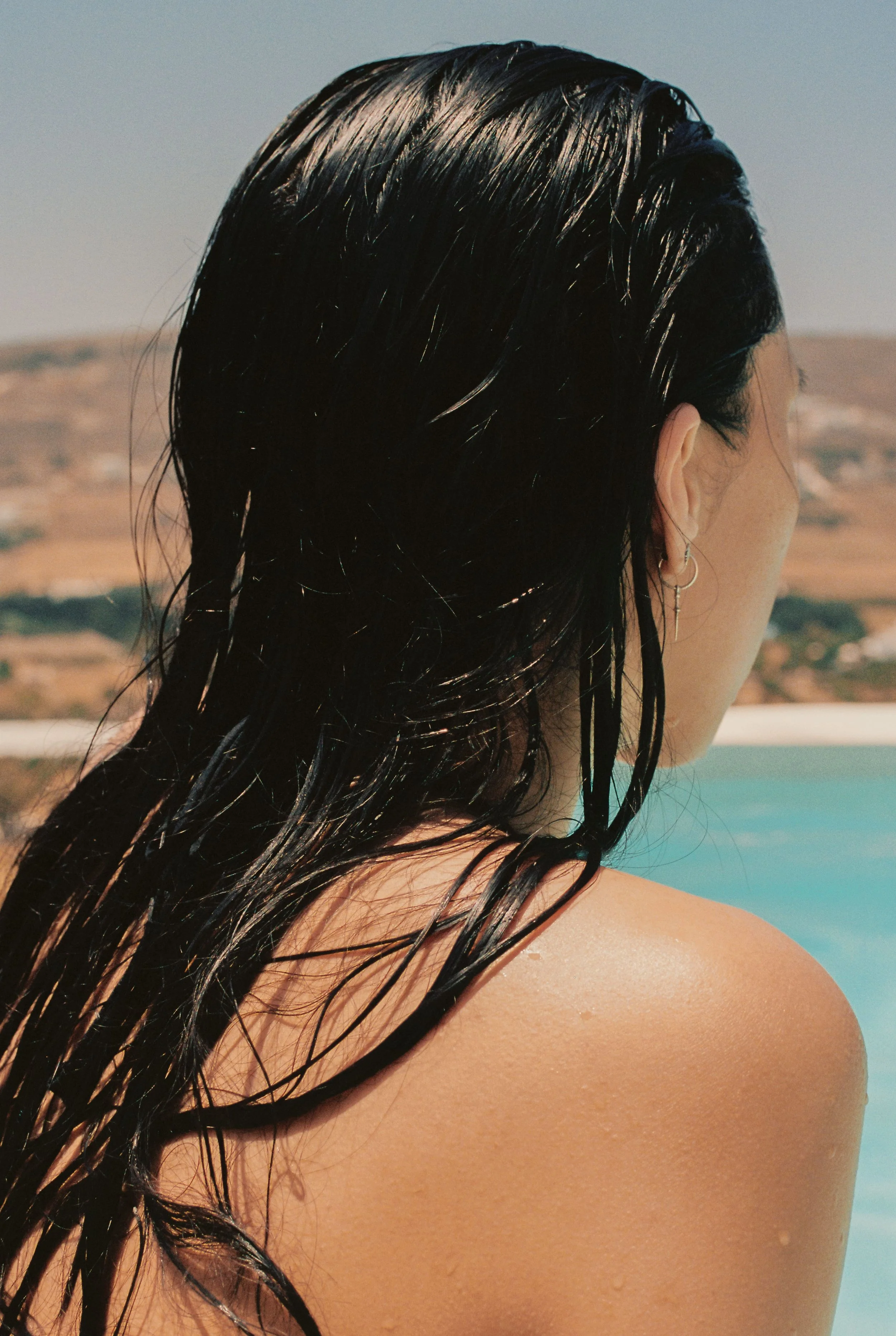 A woman with wet black hair facing away outdoors with mountains and sky in the background.