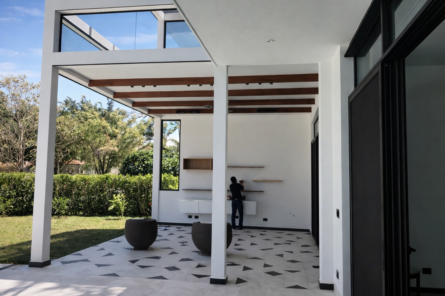 Modern outdoor patio with white walls, large glass windows, wooden ceiling beams, and black and white patterned tile floor, overlooking a garden with trees and shrubs.