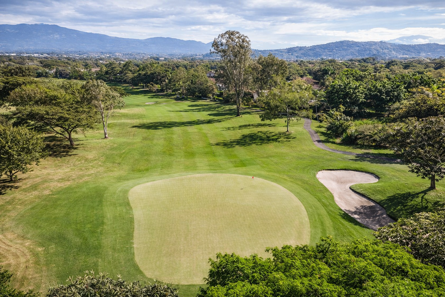 Aerial view of a golf course with a green, sand traps, and trees, surrounded by landscape including mountains in the background.