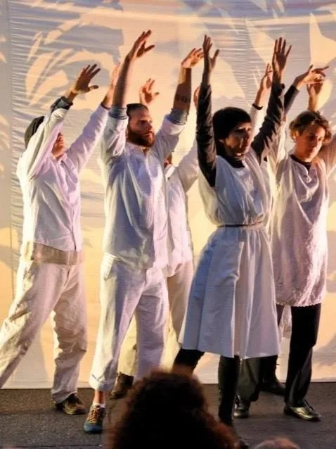 Group of five people standing on stage with arms raised, wearing light-colored clothing, in front of a backdrop with shadows and patterns.