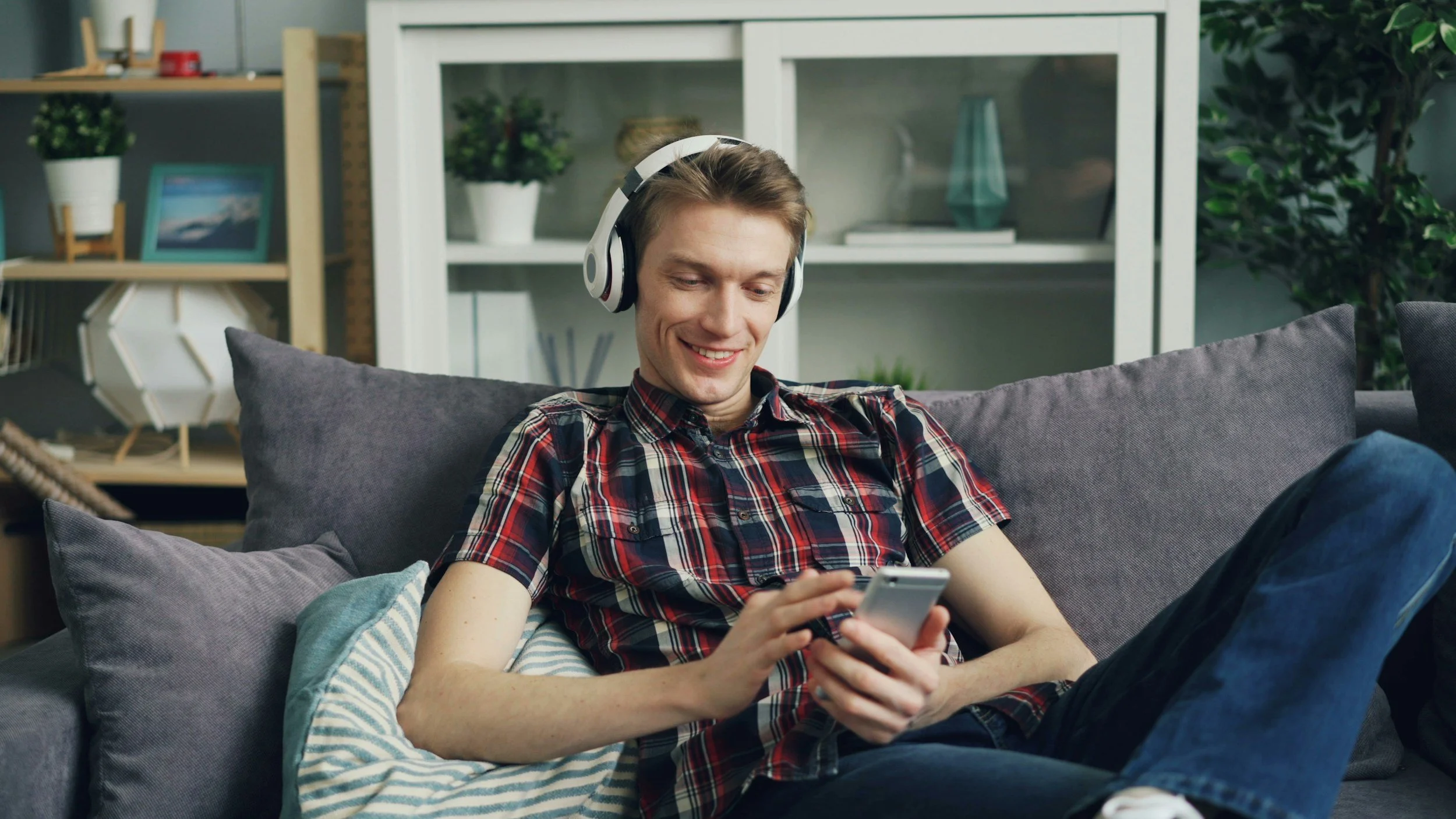 A young man sitting on a gray couch, wearing headphones and a red plaid shirt, smiling and looking at his smartphone.