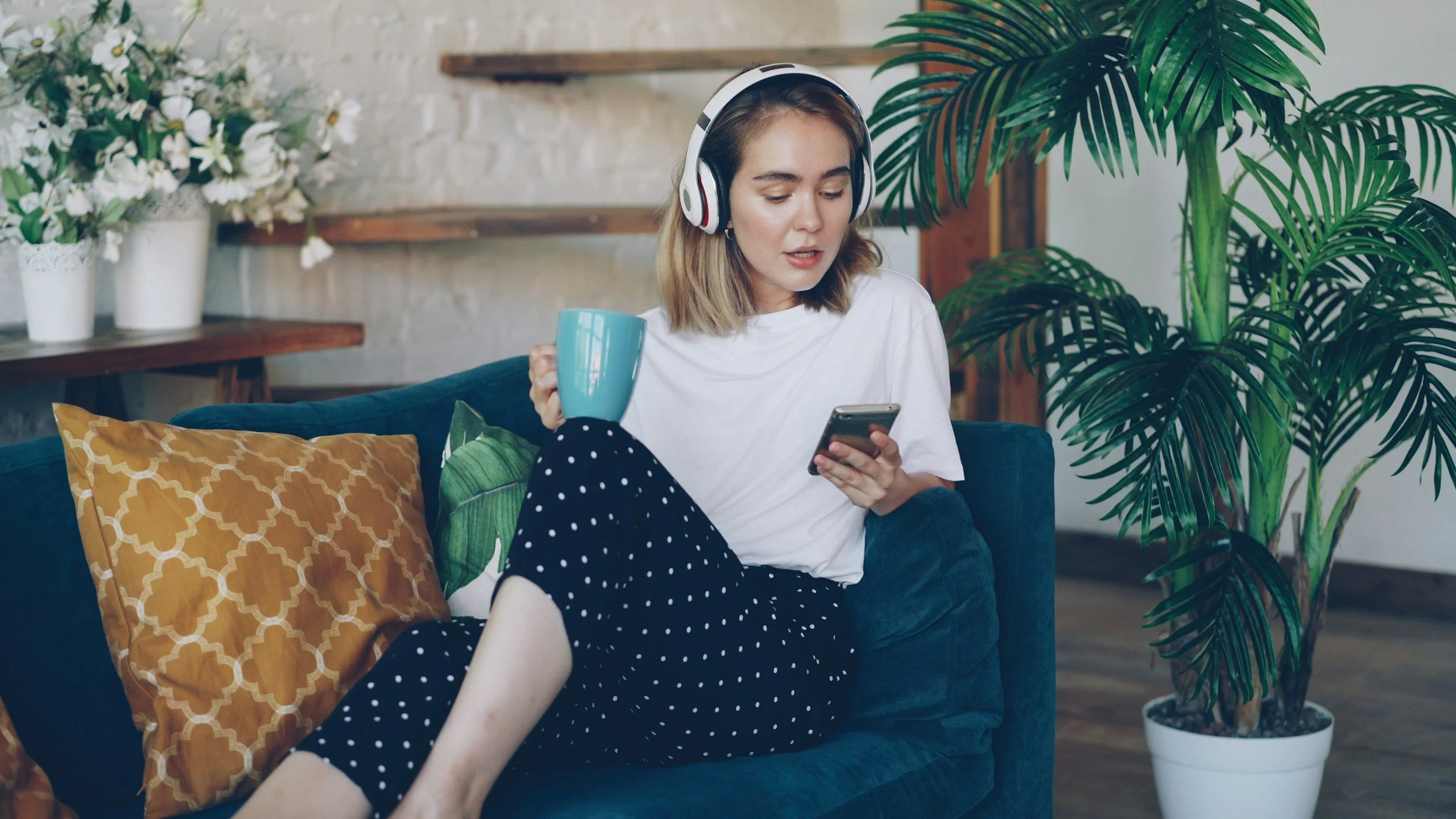 A young woman with headphones, holding a blue mug, sitting on a blue sofa, looking at her phone in a cozy room with plants and decorative pillows.